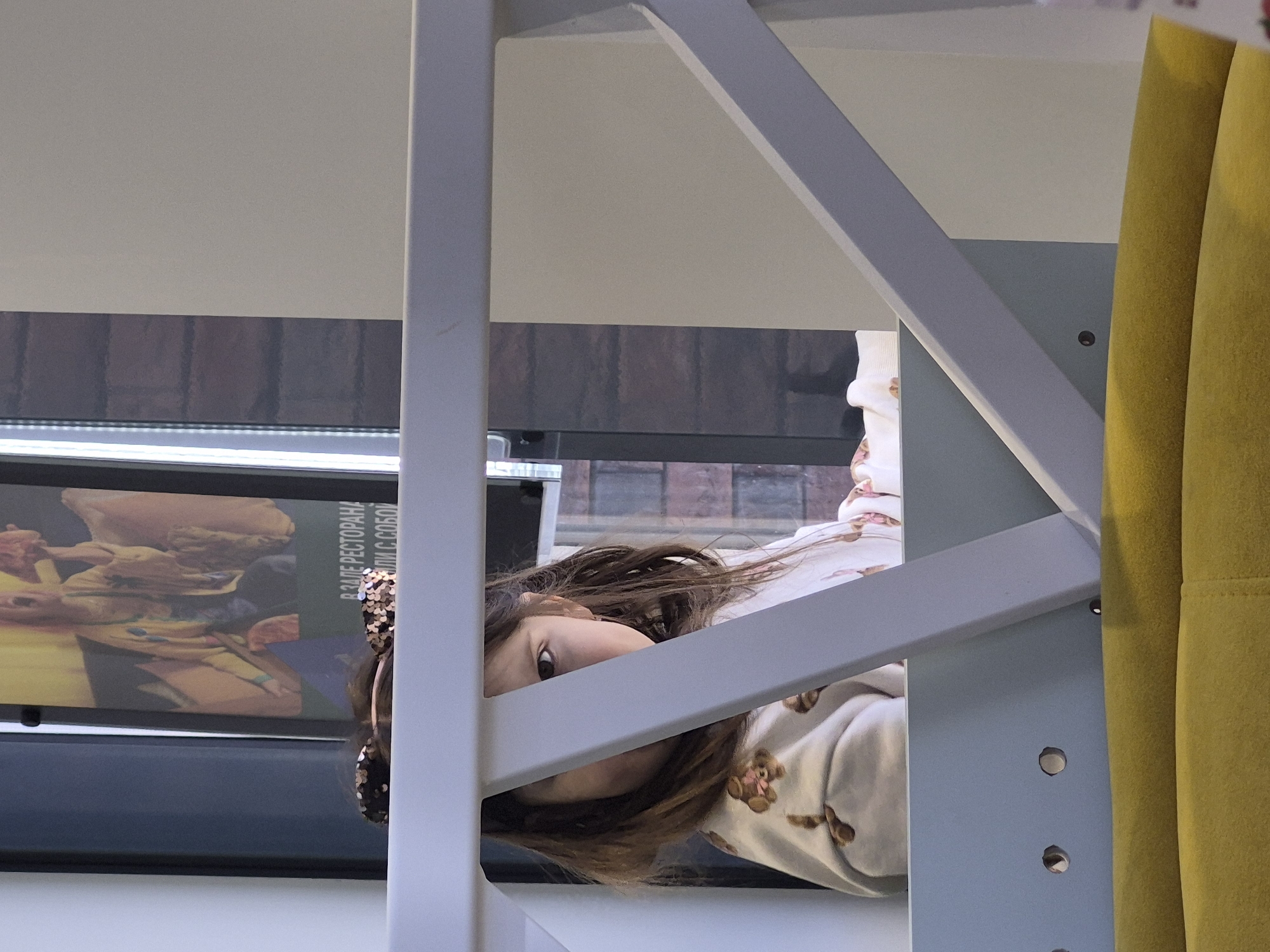 A young girl with long brown hair, wearing a headband, is peering through a white bed frame. She is wearing a light-colored pajama top with brown bear designs. The background includes a window with a view of a brick building. In front of the window, there is a sign in Russian that says "В ЗАЛЕ РЕСТОРАНА ИЛИ С СОБОИ". The location is potentially Izhevsk, Russia. A yellow curtain is visible on the right side of the frame. The girl is looking towards the viewer.