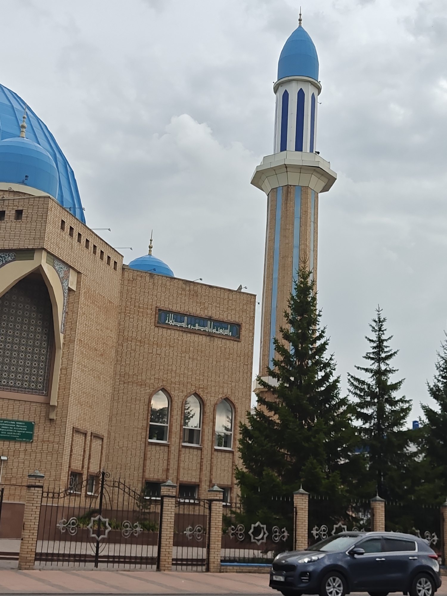 In the city of Petropavl, Kazakhstan, a large, multi-domed mosque is visible. The main structure is made of light brown brick, with ornate windows and decorative elements. A tall, slender minaret, painted with alternating blue and light brown vertical stripes, rises alongside the mosque, topped with a blue dome. Several evergreen trees are positioned in front of the minaret and the mosque. A dark blue SUV is parked on the street in the foreground, to the right of a decorative metal fence. The sky is overcast and grey, suggesting a cloudy day.