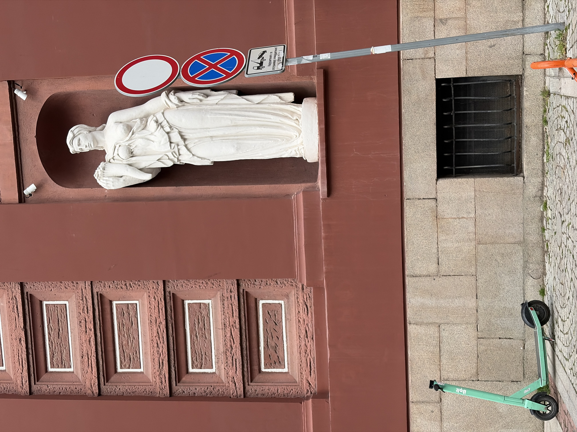 The image captures a detailed section of an urban streetscape, featuring a building facade, sidewalk, and various objects. The main subject is a white, classical-style statue of a female figure, depicted in flowing drapery, reclining within a reddish-brown niche on the building's exterior. To the left of the statue, two small white security cameras are mounted on the wall.
Above the statue, a metallic pole extends from the building, holding two prominent traffic signs: a red circular sign with a white center (likely "No Entry" or similar) and a "No Stopping" sign (red circle with blue background and a red 'X'). Below the "No Stopping" sign, another smaller sign shows a tow truck icon and Cyrillic text, which reads "Нарушителите се отстраняват принудително 0700 13 411" (Violators are forcibly removed 0700 13 411), indicating parking restrictions and a tow-away policy.
The building facade itself is a reddish-brown color, with a decorative band of recessed rectangular panels, each having a textured inner surface framed by a white outline.
On the ground level, adjacent to the building, a section of stone paving is visible. Embedded in this paving is a rectangular grated window, likely providing light or ventilation to a basement or lower level. Resting on the paving stones is a bright green electric scooter, branded "Bolt," lying on its side. The ground transitions from larger, lighter-colored paving slabs to smaller, patterned cobblestones further from the building, with small patches of grass growing between them.
The scene appears to be set during the daytime, with soft, even lighting suggesting either an overcast sky or indirect sunlight. The Cyrillic text on the sign confirms the location context of Sofia, Bulgaria. No specific activity is occurring, presenting a static snapshot of an urban environment.