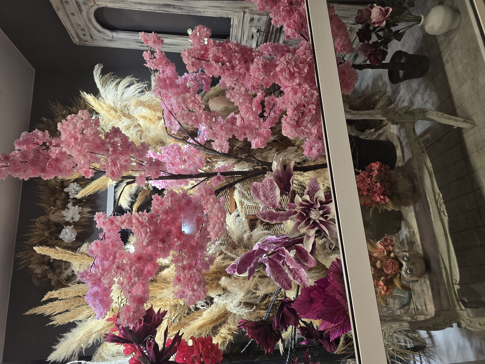 This is a close-up, eye-level shot of a floral display in a shop window in Nea Ionia, Greece. The arrangement features a dominant artificial cherry blossom tree with numerous pink blossoms, positioned in the center and extending to the right. To the left and behind the cherry blossoms, a large bouquet of dried pampas grass and wheat stalks provides a neutral, textural background. Interspersed with the pampas grass are deep magenta and purple artificial flowers with intricate petal structures, and fanned-out, dried palm leaves in a vibrant magenta.
Below the main display, a distressed, antique-style white wooden table or console is visible. On the table, several smaller decorative elements are placed. On the left, a small pink owl-shaped ceramic pot holds dried flowers and a single pink rosebud. Further to the right, a dark planter with a smaller, artificial pink flowering plant is visible. To the far right, a white vase holds a bouquet of artificial roses in shades of pink and deep red. The entire display is viewed through a clear glass window with a white horizontal band across the bottom. The shop's interior, with a gray wall and a glimpse of a white, arched decorative element on the right, is visible behind the floral arrangement. The lighting is somewhat diffused, creating soft shadows.