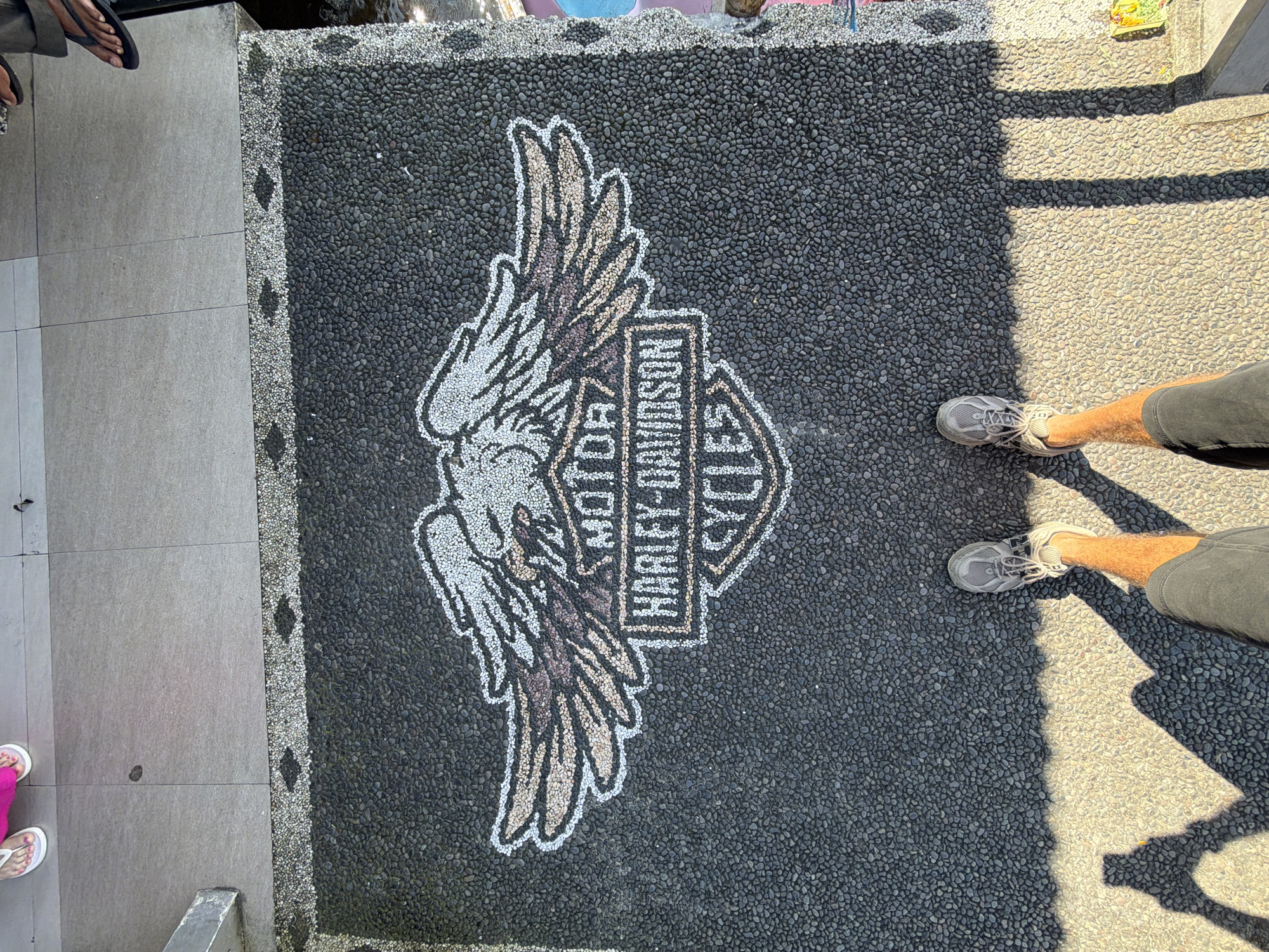 In Baturiti, Indonesia, a Harley-Davidson logo made of pebbles is embedded in a dark asphalt surface. The logo features an eagle with spread wings above a shield-shaped emblem containing the text "MOTOR HARLEY-DAVIDSON CYCLES". The logo is bordered by smaller pebbles and a concrete walkway. Two individuals are partially visible in the frame. One person, wearing flip-flops and pink clothing, stands on the walkway. Another person, wearing gray sneakers and shorts, stands on the asphalt, casting a shadow that stretches across the logo. The scene appears to be outdoors, under natural sunlight.