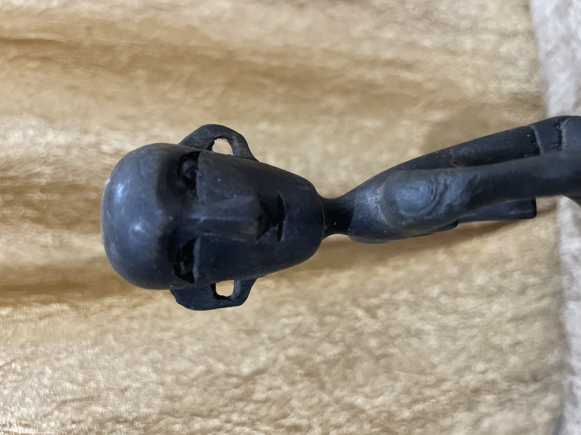 A black, carved wooden figurine of a stylized human figure is centered against a blurred, golden-brown textured background. The figurine has a prominent head with carved facial features including almond-shaped eyes, a triangular nose, and a small mouth. Large, oval-shaped ears with cutouts extend from the sides of the head. The body is elongated and thin, with visible carved lines suggesting muscles or joints. The figure appears to be in a seated or crouching pose with its legs bent. The background appears to be fabric with a soft, plush texture, possibly a draped cloth or blanket, which is out of focus, drawing attention to the figurine. The lighting is soft, casting subtle shadows that highlight the carvings on the figurine. The overall impression is that of a handcrafted artifact displayed against a neutral backdrop. The image does not provide any information or context about the city of Bhagsu, India.