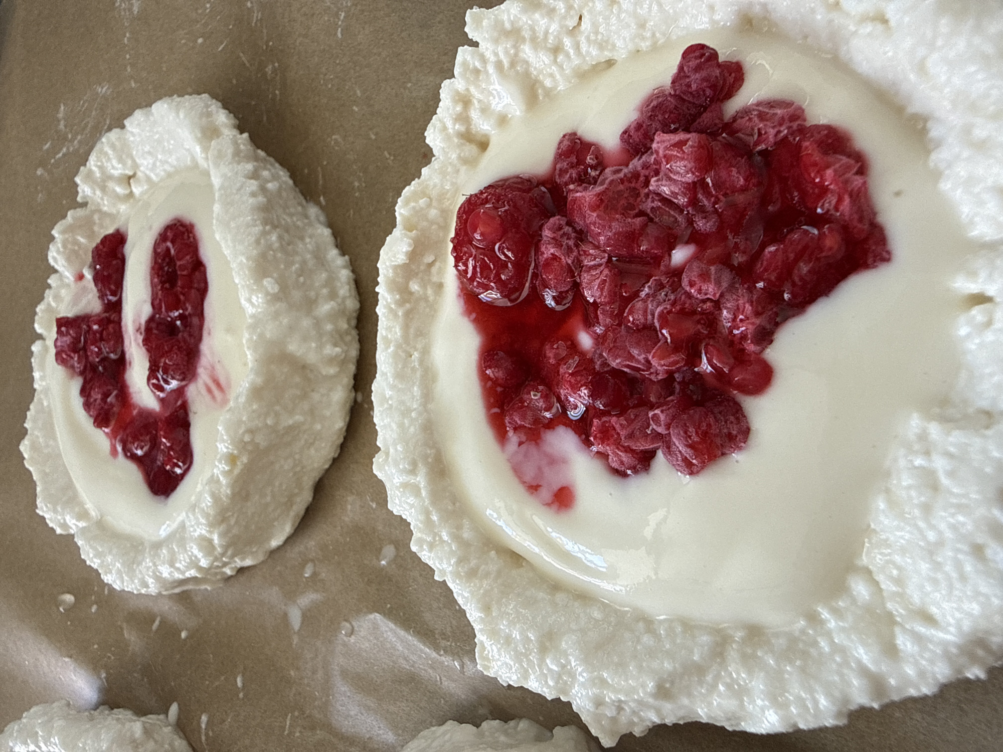 The image shows two round desserts with white, textured edges and creamy white centers topped with a cluster of red raspberries and a red sauce. The desserts are sitting on a light brown surface, likely parchment paper, which is sitting on a table. No people are visible. There is no explicit indication that this image was taken in Chaikovsky, Russia.