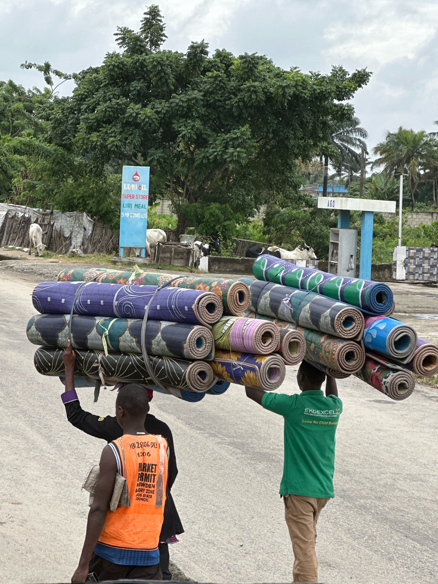 Two young men are carrying a large stack of rolled-up mats on their heads on a paved road in Badagry, Nigeria. The man in the foreground wears an orange safety vest with writing on the back that includes "MARKET PERMIT" and "BADAGRY ZONE". He is also wearing a dark purple shirt and brown pants. The man behind him wears a green polo shirt with the logo "EKOEXCEL" and "EXCELLENCE IS ON A VICTORIOUS LEARNING JOURNEY" and "Leave No Child Behind", and he is wearing brown pants. The mats are stacked high and tied together with ropes. The background shows trees, a blue sign that reads "LORIL OIL SUPER STORE LURI MEAL", and a white structure with "AGO" on it, which is likely a gas station. There are also some cows in the background. The sky is overcast.