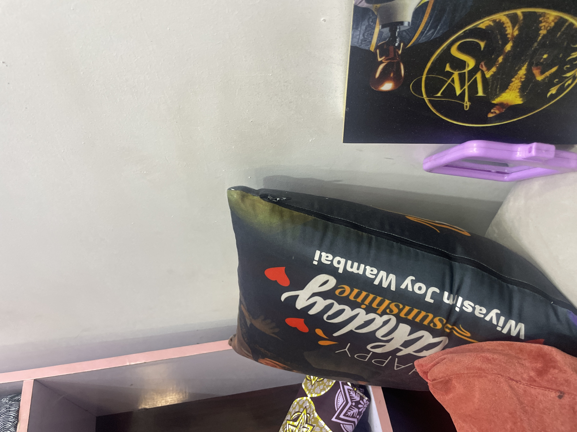 This image shows a pillow with the words "Happy Birthday Sunshine Wiyasin Joy Wambai" printed on it in white and yellow lettering. The pillow is black with an abstract design. There are also two small red hearts and some orange and yellow shapes on the pillow. The pillow is resting on a shelf, and a pink object is visible behind it. A framed picture is on the wall above the pillow. The picture is dark with a yellow oval logo in the center. The overall scene appears to be indoors, possibly in a home or a shop. The image is taken from a slightly tilted angle.