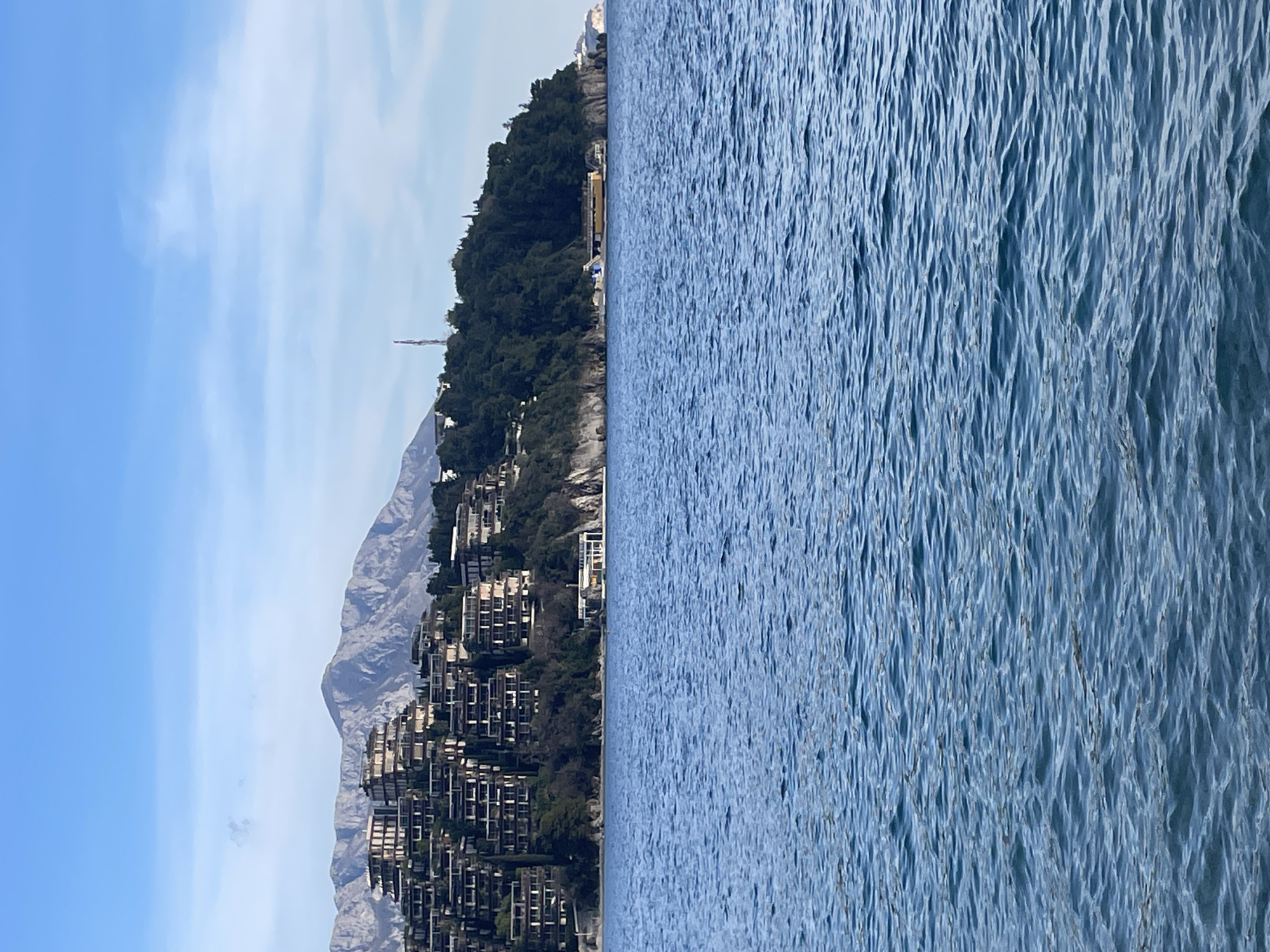 The image captures a coastal landscape view of the Zavala Peninsula in Budva, Montenegro. The foreground is composed entirely of rippled blue seawater. The midground features a verdant coastline with a prominent cluster of multi-story, terraced buildings, identified as Dukley Gardens, integrated into the green hillside on the left. These structures display a modern design with numerous balconies. Further along the coastline to the right, a smaller white multi-level building with a flat roof is situated on a rocky outcrop at the water's edge. Beyond this, a long, yellowish-orange floating structure, potentially a pontoon or similar marine facility, is visible offshore near the base of the headland. A tall, slender red and white cellular tower stands atop the peninsula's ridge. The background is dominated by a rugged, grey mountain range, characterized by visible peaks and valleys. The sky is clear light blue with scattered, faint white clouds. No people, specific actions, or interactions are discernible within the frame.