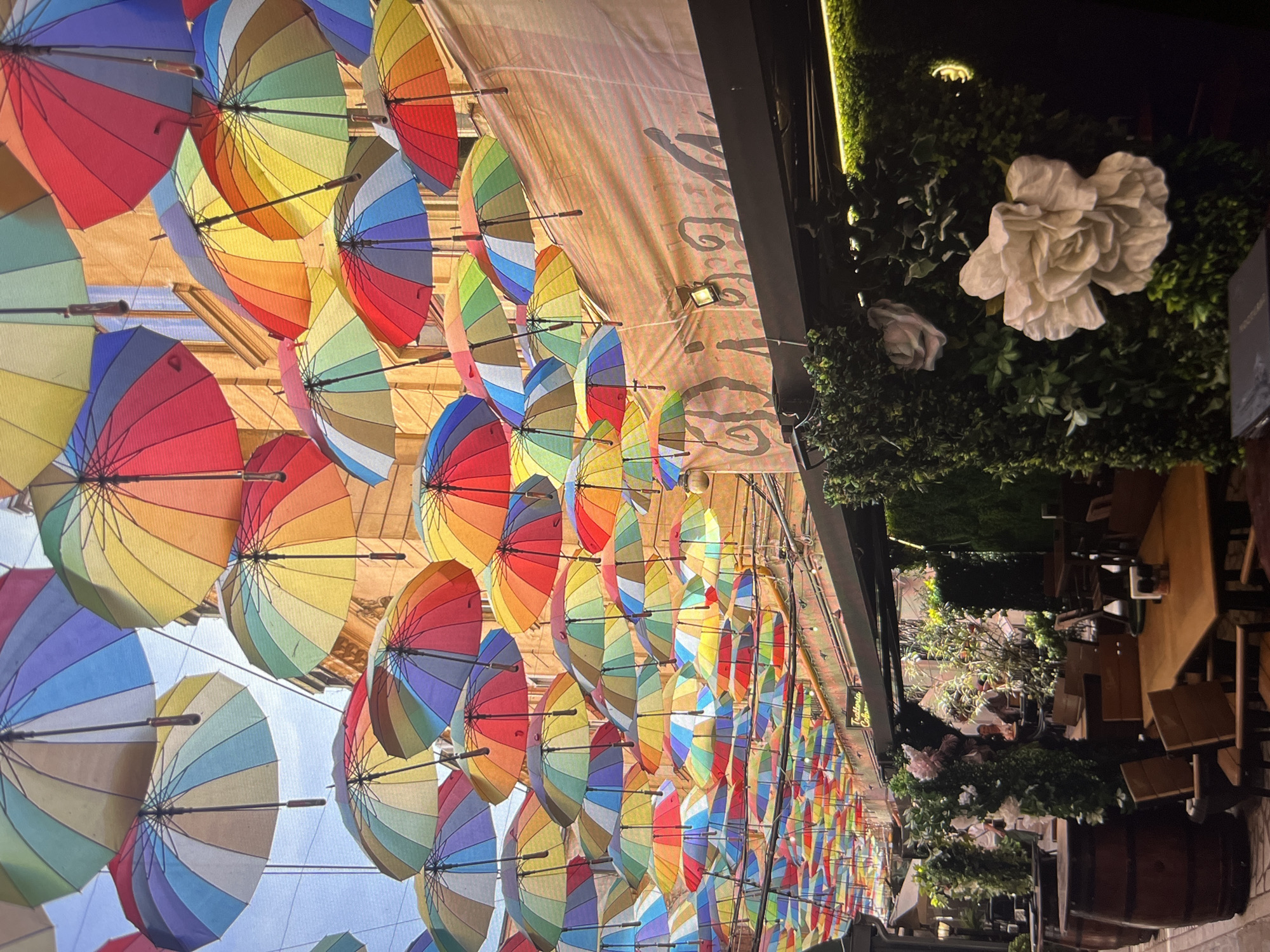 The image shows a street in an unknown city, featuring an outdoor dining area decorated with numerous rainbow-colored umbrellas suspended overhead. The umbrellas provide shade to tables and chairs set up for patrons. Greenery, including flowers, adorn the side of the building, adding a touch of nature to the urban environment. Tables are set with dining items suggesting a restaurant or cafe setting. The scene appears to be taken at dusk or night, illuminated by artificial lighting.