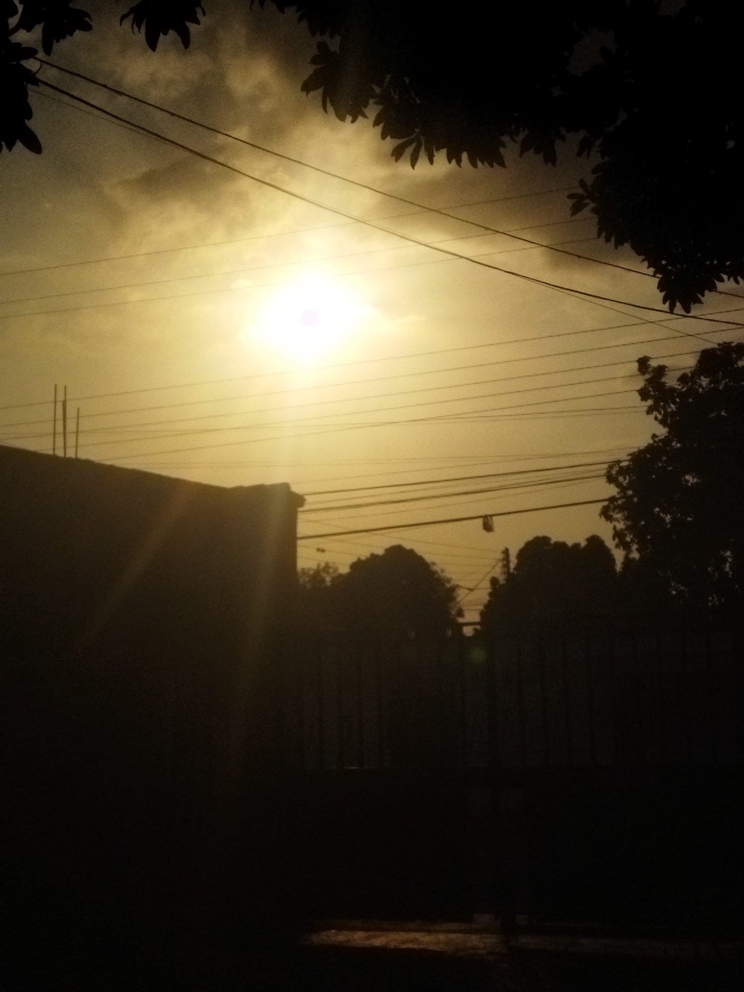 The image shows a hazy or cloudy sky in Unknown, Unknown, with a bright sun obscured by the atmospheric conditions. Power lines are visible across the sky. Silhouetted against the bright sky are trees, the top of a building with vertical protrusions, and a fence. The foreground is dark, with a glimpse of the ground.