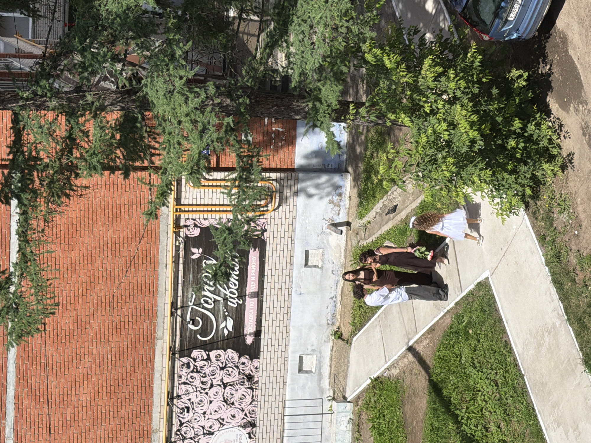 In Chaikovsky, Russia, on a sunny day, a group of four people stand on a concrete path in front of a brick building with a floral shop sign. The sign, featuring pink roses and text, is mounted on the building's facade. A young girl in a white dress and a white hair accessory stands to the right of the group, facing away from the camera, holding a bouquet of flowers. To her left, three individuals are gathered on the path: a man in a white shirt and grey pants, a woman in a dark brown dress, and another woman in a dark dress. The man appears to be speaking. The area is surrounded by green grass and a large tree with lush foliage casts shadows on the ground. A blue car is partially visible to the right of the frame. The building exhibits signs of wear, with peeling paint on the lower section.