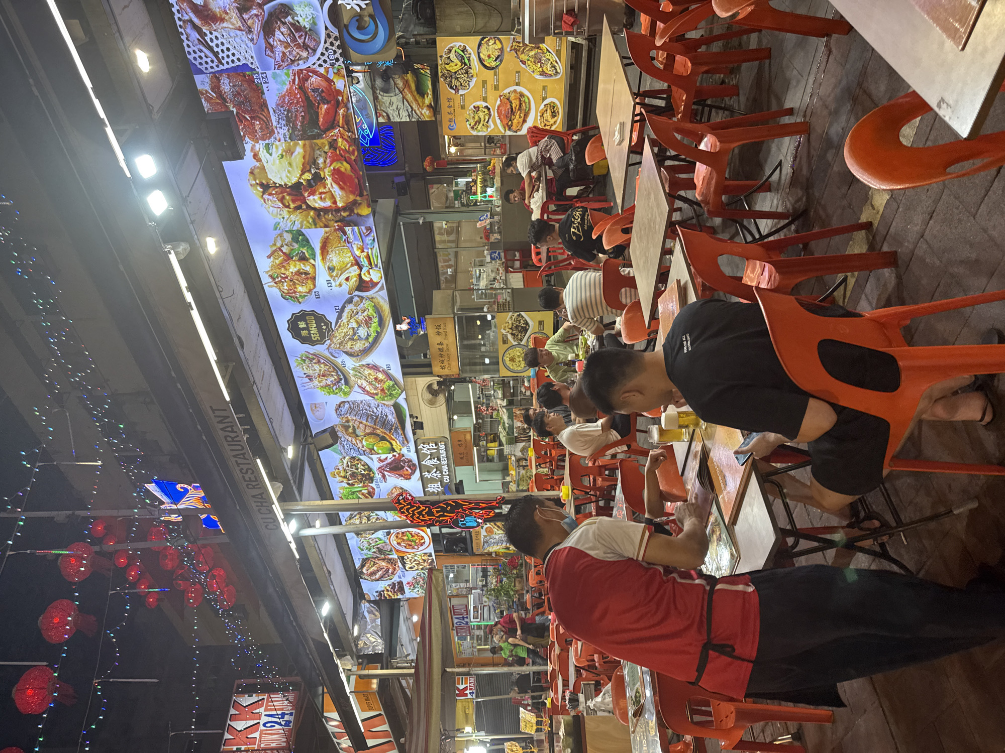 This vibrant nighttime scene depicts an active outdoor dining area, likely a hawker center or street food establishment, in Kuala Lumpur, Malaysia. Numerous patrons, predominantly male, are seated at simple wooden tables with bright orange plastic chairs. They are engaged in dining and social interaction, with some individuals looking at their mobile phones. Glasses of beer are visible on several tables, suggesting a casual and convivial atmosphere.
The setting is illuminated by bright overhead fluorescent lights, casting a strong glow on the seating area. A prominent feature is the series of large, brightly lit digital menu screens displaying an array of appetizing seafood dishes such as grilled fish, crabs, and lobsters, along with their prices. Festive red lanterns and strings of colorful fairy lights hang from the awning, adding to the lively ambiance. A distinctive neon sign in the shape of a red and blue creature, possibly a dragon, is also visible.
Visible text includes "CU CHA RESTAURANT" and "粗茶食馆" (its Chinese name), indicating the name of the eatery. Other signs read "海鮮 SEAFOOD," "松城炒粿条炒饭 Char Kuey Teow Fried Rice," and "BUKAPA JA," suggesting local specialties. In the background, "KK SUPER MART" and "A24JAM" (24 hours) signs further confirm an urban, commercial setting. The image captures the dynamic energy of a popular dining spot after dark.
