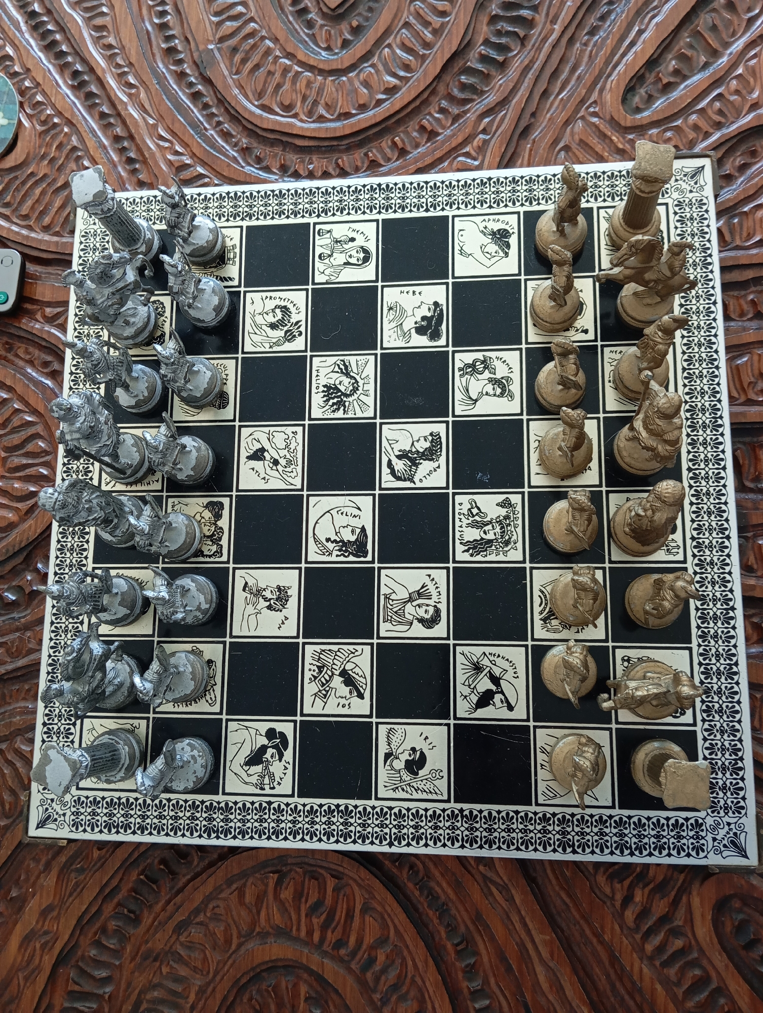 An overhead shot captures a unique, elaborately designed chessboard set up on a dark, intricately carved wooden table. The chessboard features alternating black and white squares, with many of the white squares adorned with black-and-white illustrations of figures from Greek mythology, each labeled with their name.
Two distinct sets of chess pieces are arranged on the board, appearing to be in their initial game positions. One set is a silver-colored metallic finish, positioned on the left side of the board. The other set, on the right, has a gold or bronze-toned metallic finish. The pieces themselves are highly detailed, figurative sculptures representing various mythological characters rather than traditional chess figures.
Visible text labels on the white squares include: "THEMIS", "PROMETHEUS", "APHRODITE", "HEBE", "HELIOS", "HERMES", "ATLAS", "APOLLO", "CELINI", "DIONYSUS", "PAN", "ARTEMIS", "HEPHAESTUS", "SATYR", and "IRIS". Partially visible text includes "ACHILLE" and "105".
The chessboard itself has a decorative border with repetitive black-and-white patterns. The rich, dark wood grain and carving of the table beneath add a warm, traditional feel to the setting. A small, light gray electronic device with a green power button is partially visible in the upper left corner of the frame. The scene suggests a ready-to-play game or a decorative display of a themed chess set, likely indoors. The lighting indicates daytime.