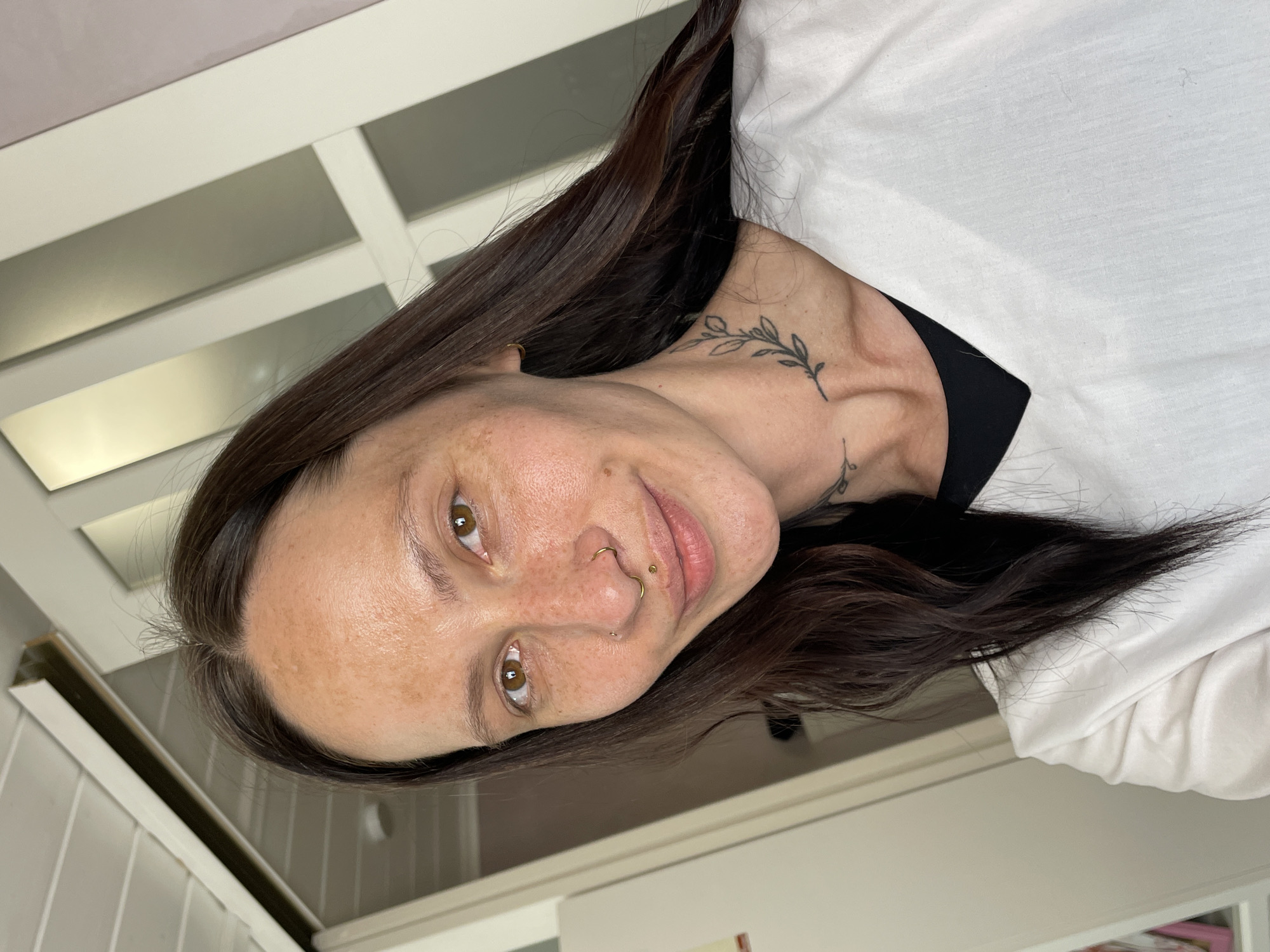 A head-and-shoulders shot depicts a fair-skinned individual with brown hair and eyes. She has freckles on her face and piercings in her nose and lip. The person is wearing a white top over a black garment. A leaf-like tattoo is visible on their neck. The background consists of white panelling. It is not possible to ascertain the location as Izhevsk, Russia, based on the visual information provided.
