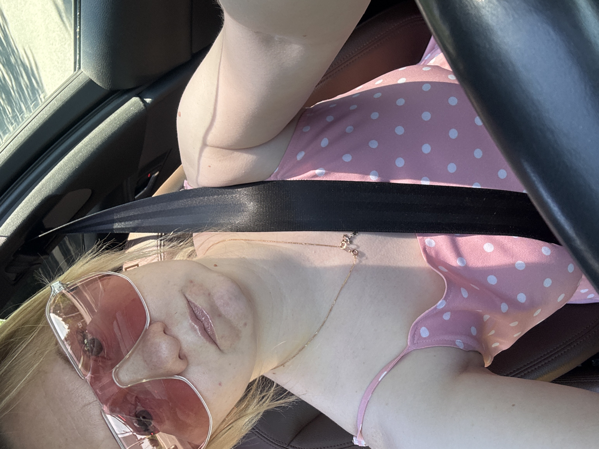 A fair-skinned woman wearing a pink polka-dot dress and rose-tinted sunglasses is seated in a car. She has a seatbelt fastened across her chest. A gold chain necklace is visible. The car's interior appears to be brown leather. The location in the city of Chaikovsky, Russia is not discernible from the image.
