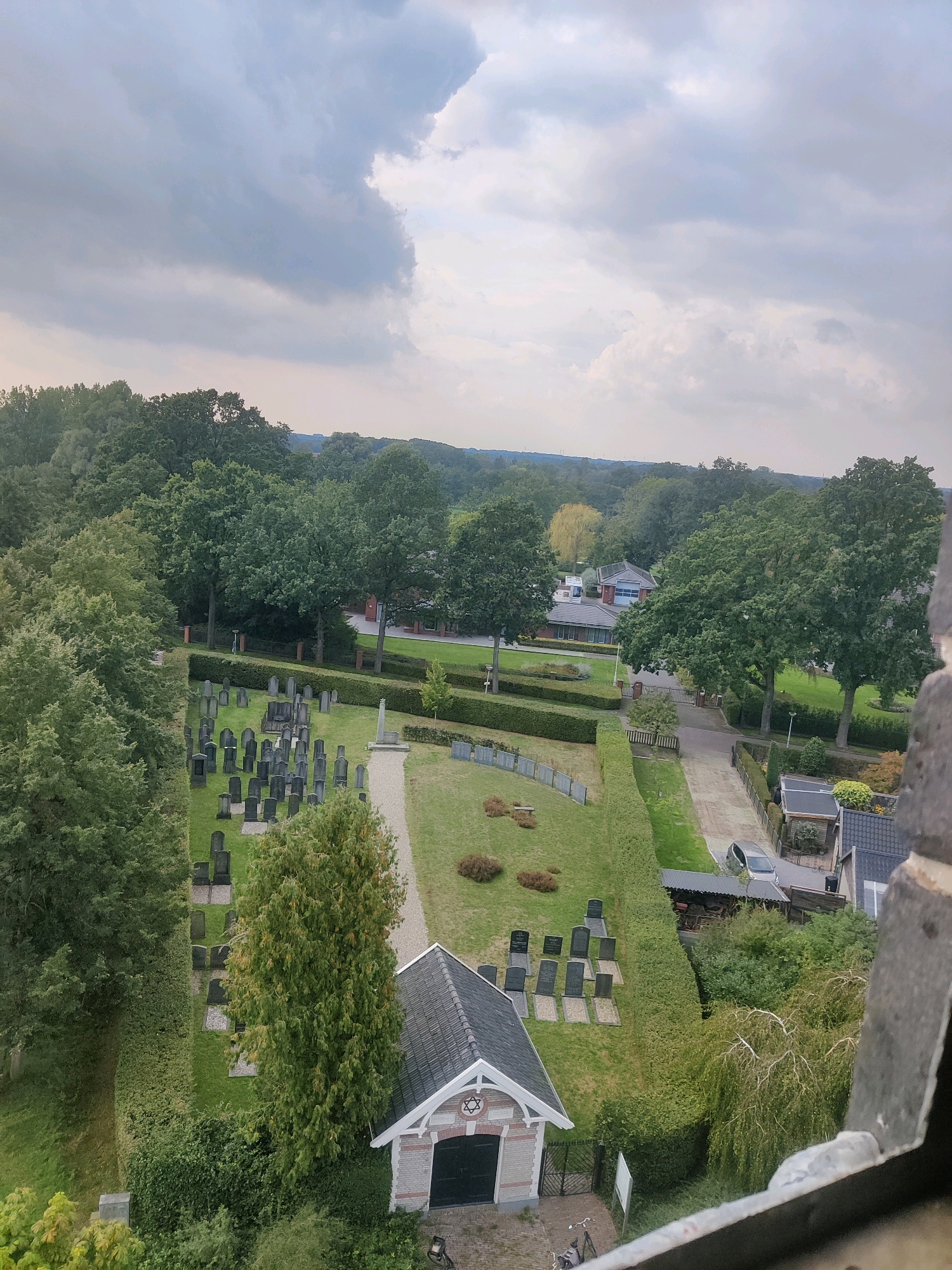 An aerial view, captured on a cloudy day, shows a cemetery in Winterswijk, Netherlands. The cemetery is characterized by rows of headstones of varying sizes and shapes, set against a backdrop of lush green grass and manicured hedges. A narrow gravel path winds through the cemetery, leading to a small building with a gabled roof, adorned with a Star of David symbol above its entrance. Adjacent to the cemetery, a residential area with houses and a street is visible, with several parked cars. The scene is framed by the textured stone of what appears to be a tower or elevated viewpoint in the foreground.
