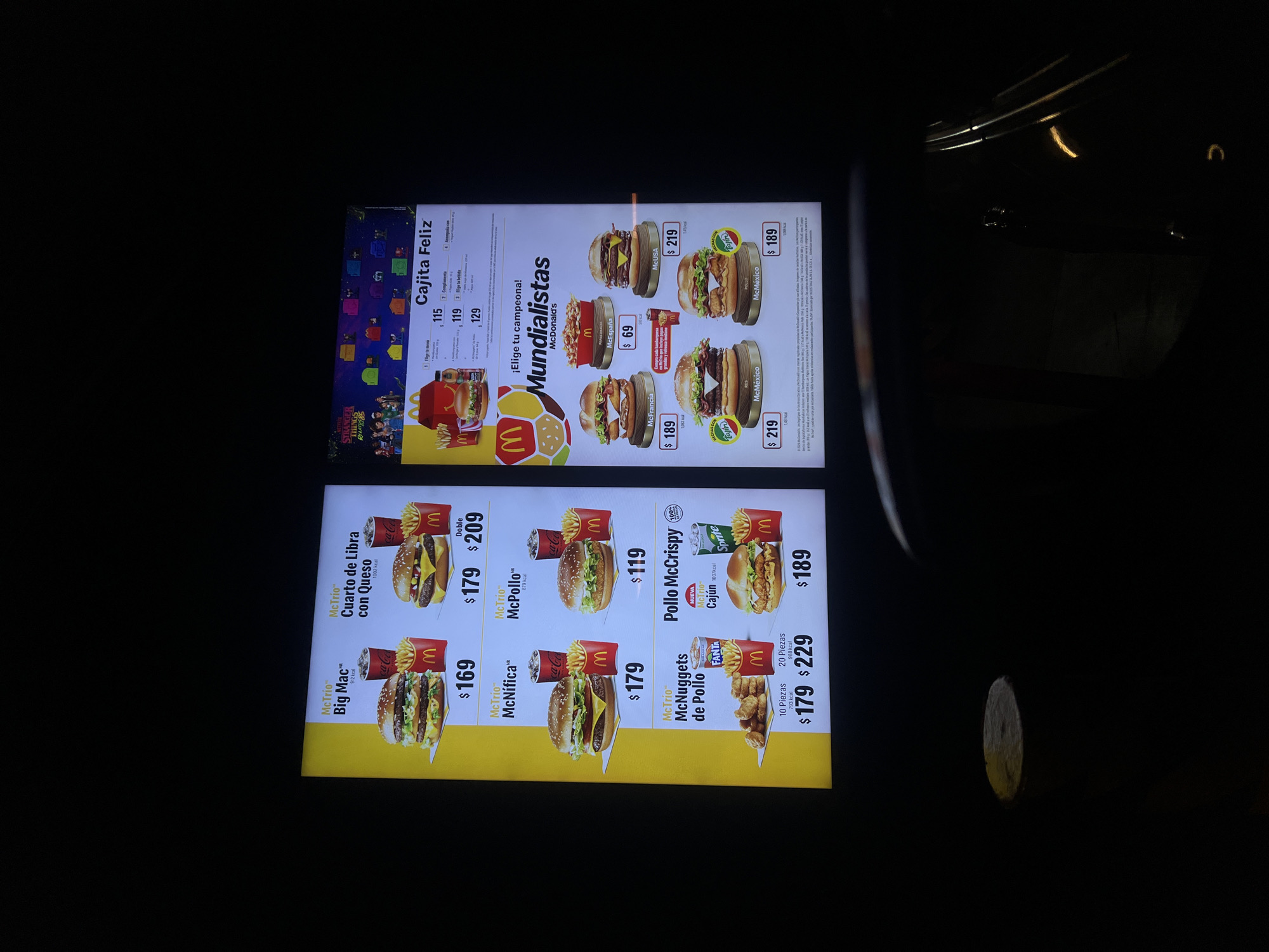 The image shows a close-up shot of two illuminated McDonald's menu boards, likely viewed from inside a car. The upper menu board advertises "Mundialistas McDonald's" themed burgers and the "Cajita Feliz" (Happy Meal). The lower menu board features standard McDonald's menu items like the Big Mac, Quarter Pounder with Cheese, McPollo, McNifica, McNuggets, and Pollo McCrispy, along with their prices in Mexican pesos. The presence of Spanish text and the monetary values strongly suggest this is a McDonald's location in Mexico, and based on the product names in the menu in Mexico City, Mexico.