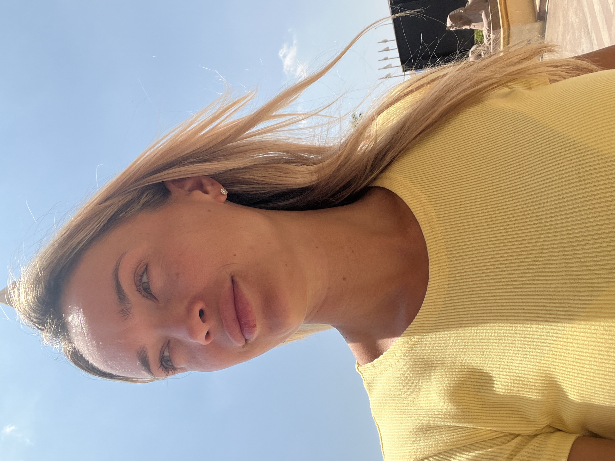 A woman with blonde hair is looking to the left, with her head tilted slightly up. She is wearing a yellow ribbed t-shirt. The background is a clear blue sky with a few small clouds. In the distance, there are flags and what appear to be sphinx statues. The lighting suggests it is daytime and sunny. This scene could be in Sharm El Sheikh, Egypt, given the presence of sphinx-like statues.