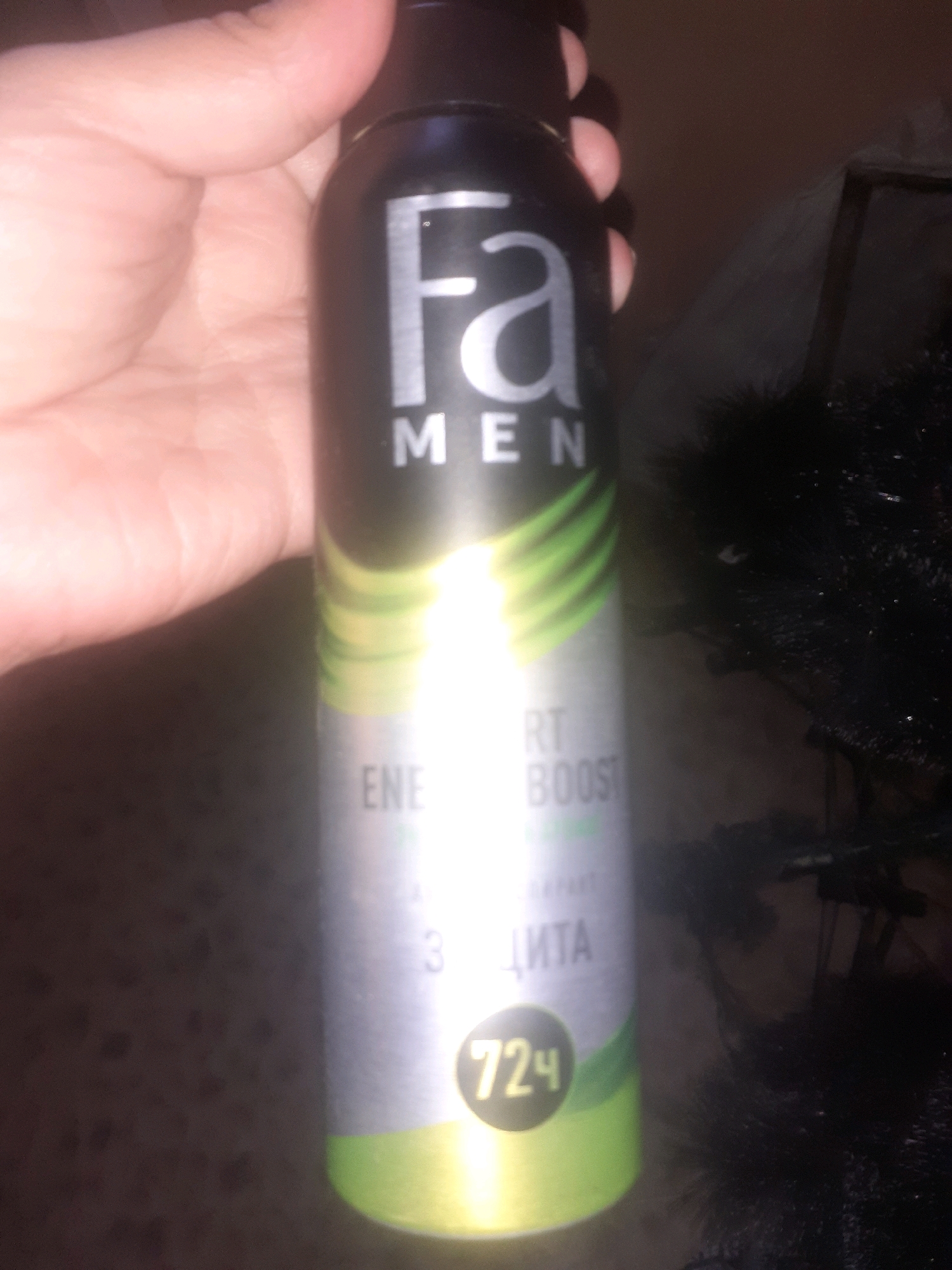 A hand holds a black aerosol can with a silver label. The label features the "Fa" logo in large silver letters, followed by "MEN" in smaller silver letters. Below "MEN," green abstract lines extend down the can. Further down the label, partially obscured by a bright light flare, are the words "ENER" and "RT BOOST." Below that, the Cyrillic word "ЗАЩИТА" (protection) is visible, and at the bottom of the can, inside a yellow circle, is the number "72Ч" (72 hours). The background is dark and out of focus, with hints of a dark, possibly artificial, plant-like object to the right. The scene is not recognizable as being in the city of Semey, Kazakhstan, or any specific urban environment.