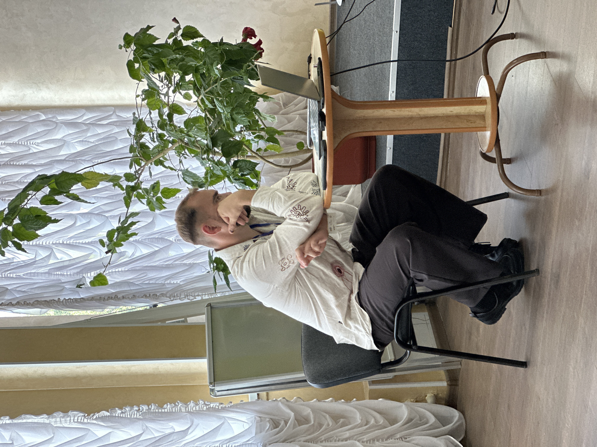 The image depicts a man sitting at a table with a laptop in Myronivka, Ukraine. He is wearing a white shirt with embroidered patterns and dark trousers. The man is leaning back in his chair, with his arms crossed and his hand near his mouth, suggesting a pensive or tired mood. A large potted plant with green leaves and red flowers is positioned to the left of the table, partially obscuring the man and the laptop. The scene appears to be indoors, with a white, textured wall and sheer curtains visible in the background. A wooden table with a distinctive curved base supports the laptop. The overall lighting suggests it could be daytime. No visible text is present in the image.