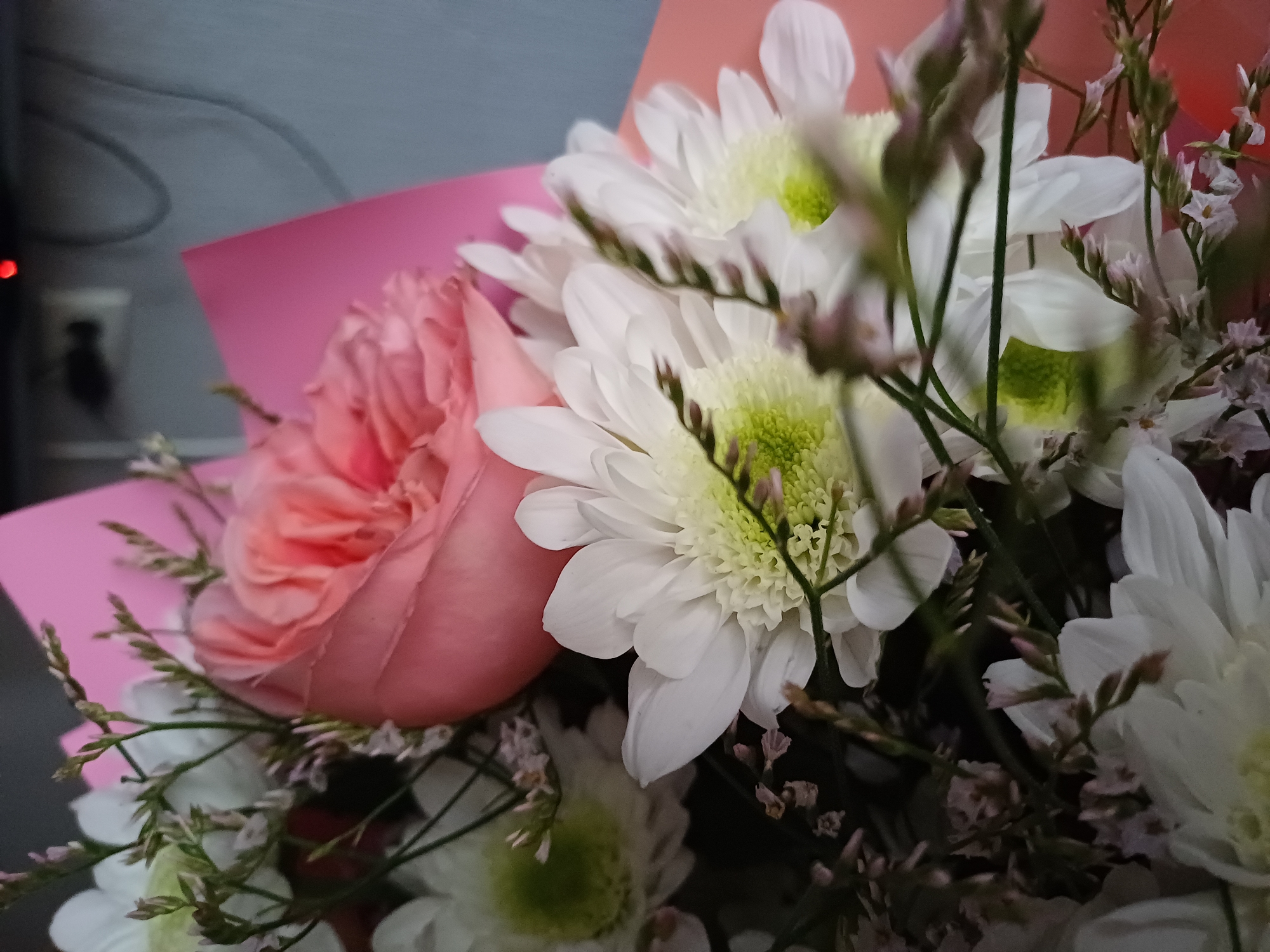 A close-up shot of a bouquet of flowers, featuring a prominent pink rose and several white chrysanthemums. The flowers are arranged with delicate green stems and smaller, dried flowers. The bouquet is partially wrapped in pink and peach paper. The background is blurred, showing hints of a grey wall and what appears to be electronic equipment, possibly a monitor with a red indicator light. The lighting is dim, creating a soft focus and emphasizing the textures of the petals and foliage.