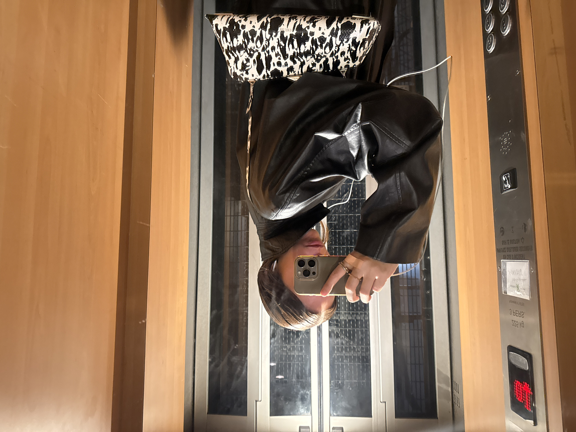 A selfie taken in an elevator shows a person in a black leather jacket holding an iPhone. A black-and-white patterned bag is above their head. The elevator's control panel is on the right, and wooden panels are on either side. The reflection in the elevator doors indicates the location is possibly in Paris, France, based on the architecture and style.