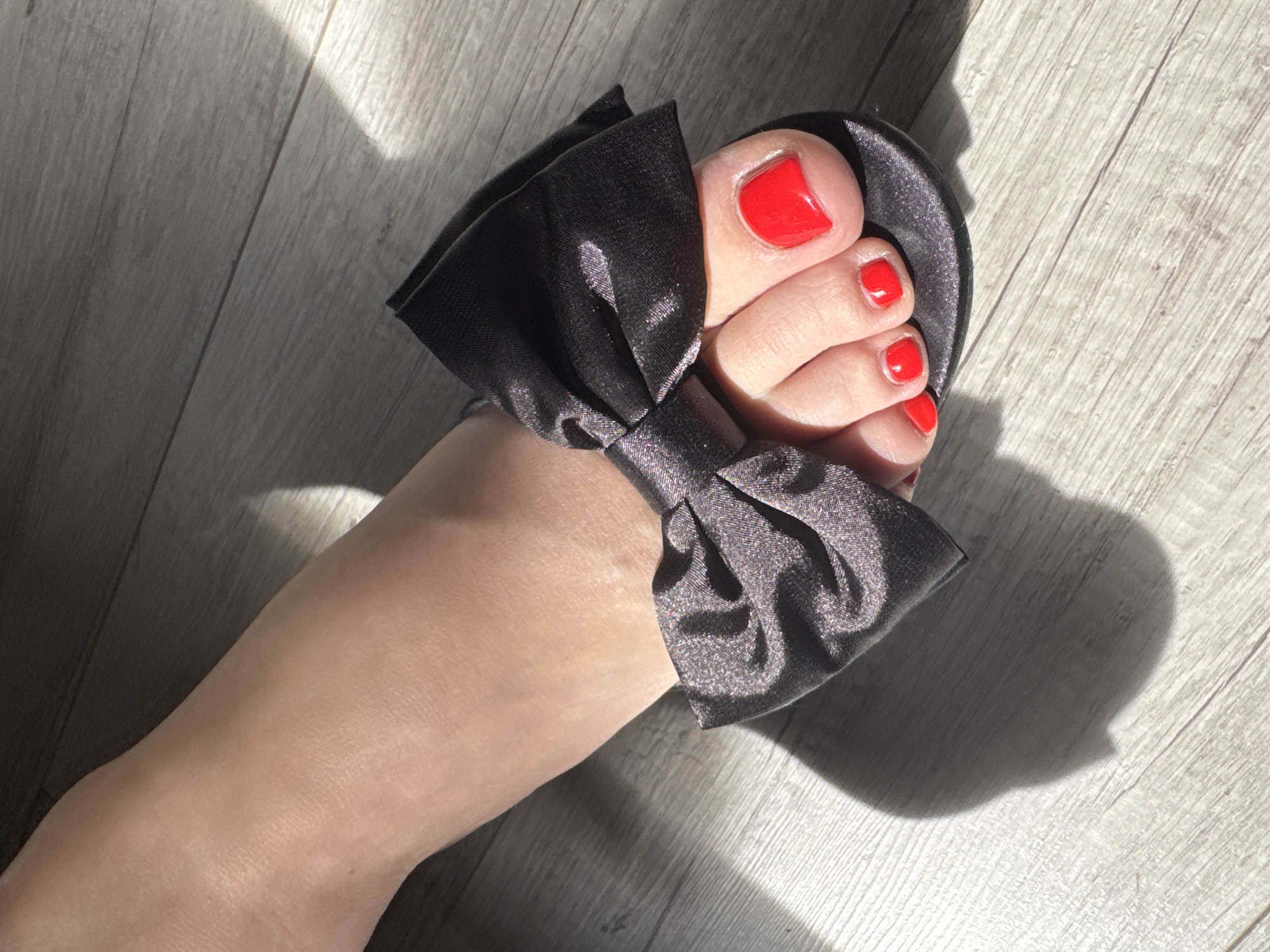 This image presents a close-up, high-angle view of a person's foot, adorned with a bright red pedicure, resting on a light-toned wooden floor. The foot is wearing a black slipper or slide, characterized by a large, satin black bow covering the top of the foot. Intense natural light, likely sunlight, casts distinct shadows on the floor and the sides of the foot, indicating it is daytime and sunny. The flooring features light-colored wooden planks with a subtle grain pattern. The scene is static, with no specific activity beyond the foot being positioned for the photograph. No text is visible in the image. The setting is an indoor space, located in Moscow, Russia.