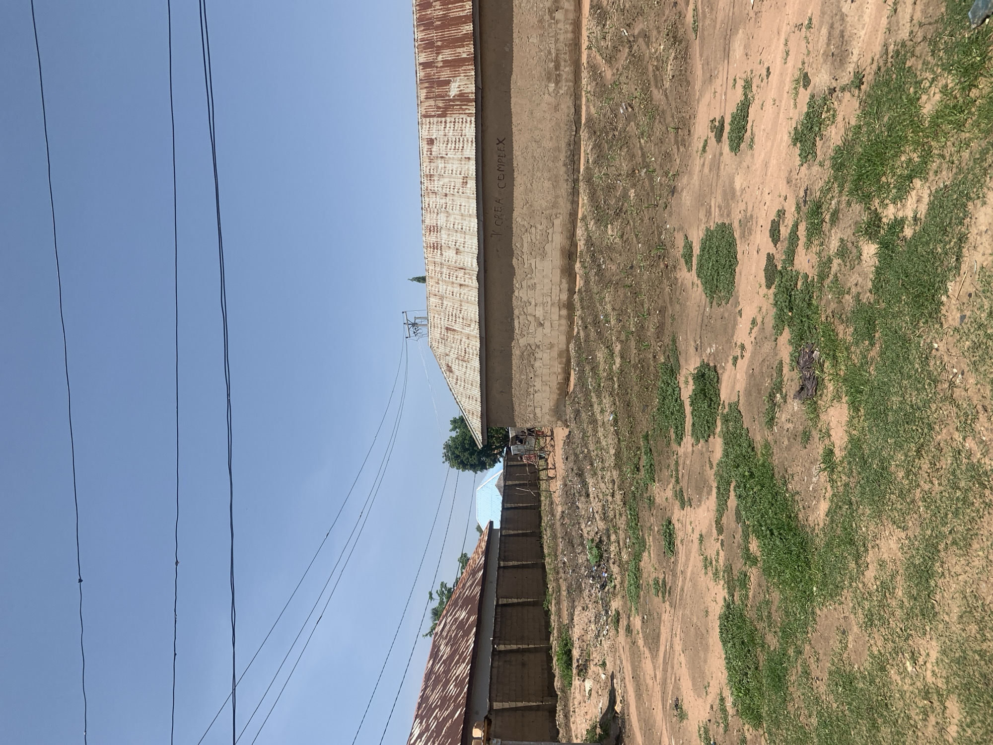 The image depicts a street scene in Jauro Yinu, Nigeria, characterized by a dirt road with sparse patches of green grass and some visible debris. Several buildings with corrugated iron roofs, some showing signs of rust, line the street. The walls of these buildings are constructed from what appears to be mud bricks. One building in the foreground has the words "Y.CREA COMPLEX" scrawled in black lettering on its upper wall. In the middle ground, two bicycles are visible, one of which is attached to a small cart. Two individuals, one appearing to be a child, are near the bicycles. Overhead, multiple electrical wires crisscross the clear blue sky, supported by poles visible in the distance. The overall environment suggests a rural or semi-urban setting.