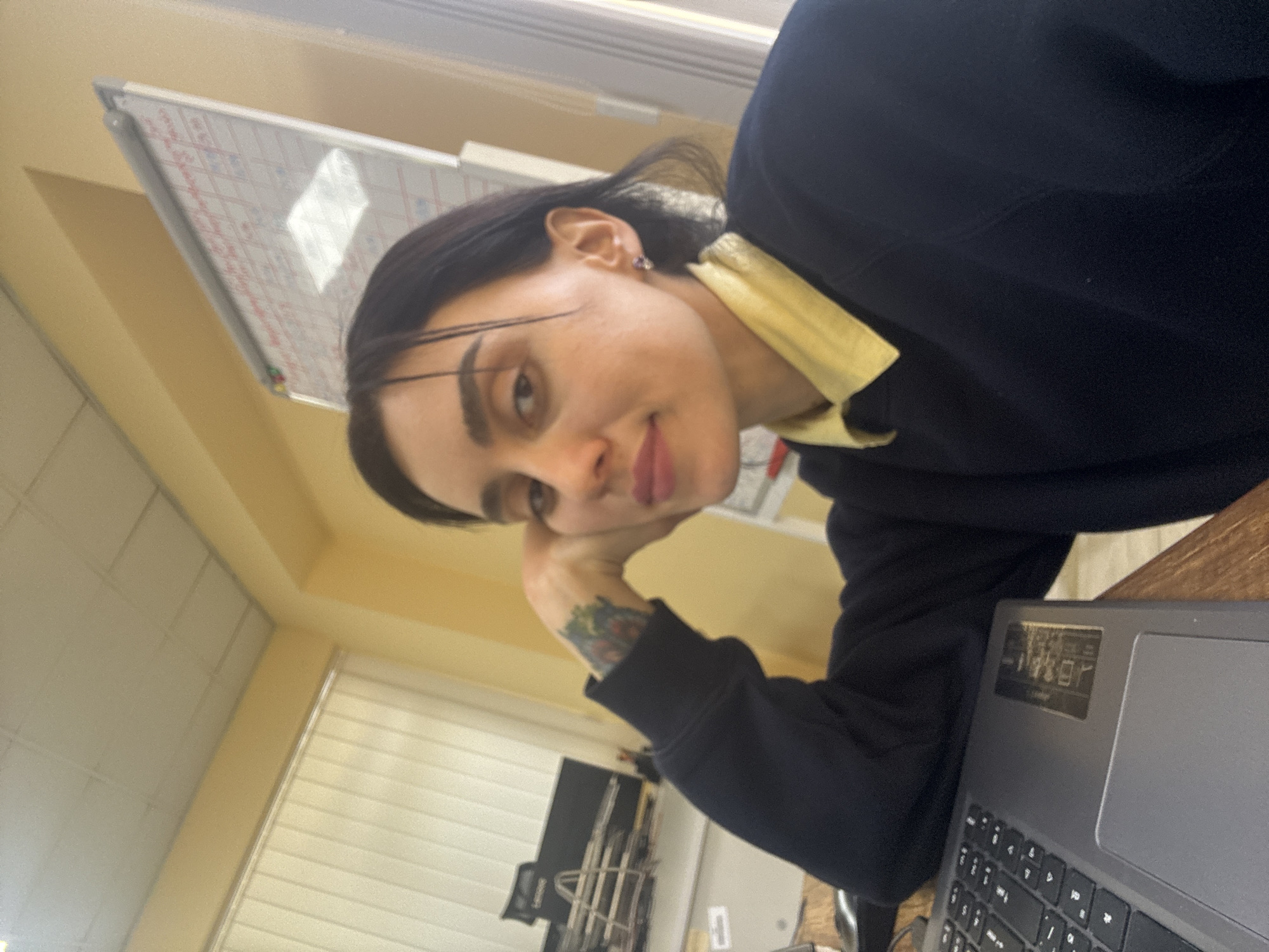 A young woman with dark hair and fair skin is resting her chin on her hand while sitting at a desk with a laptop. The woman is wearing a black sweatshirt over a yellow collared shirt, and she has a tattoo on her left wrist. A whiteboard with markings hangs on the wall behind her. In the background, there is a window with blinds and a view of what appears to be construction scaffolding outside. Given the architectural style and available information, the image could potentially be located in Tbilisi, Georgia, but this is not definitively confirmed.