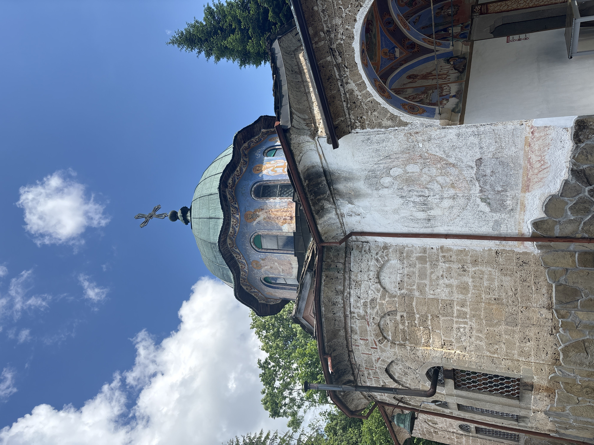 The image captures a detailed view of a historic Eastern Orthodox monastery or church, situated in what is identified as the city of Charkovo, Bulgaria, under a bright blue sky with scattered white clouds. The central feature is a large, weathered green dome topped with a metallic Orthodox cross. Below the dome, a cylindrical drum is adorned with faded but discernable frescoes depicting religious figures against a predominantly blue background, featuring two arched windows.

The building's architecture showcases a combination of materials. The lower left section of the structure is constructed from light-colored, irregular stone blocks, forming several arched recesses and one window fitted with an intricate metal grille. A small, dark green bell hangs from a metal support frame on the far left. A dark metal downspout runs vertically down this stone wall. The wall transitions to a lighter, weathered plastered section on the right, displaying faint traces of paint or frescoes.

On the far right, an arched entrance or passage is visible, its interior ceiling and walls richly decorated with colorful frescoes depicting various religious scenes and figures, primarily in blue, red, gold, and white hues. A small, light-colored table is positioned within this archway. Dark brown gutters are visible along the rooflines and various architectural elements. Lush green trees, including a tall coniferous tree on the right and deciduous foliage on the left, frame the building against the backdrop of the clear, partly cloudy sky. No people are visible in the scene.