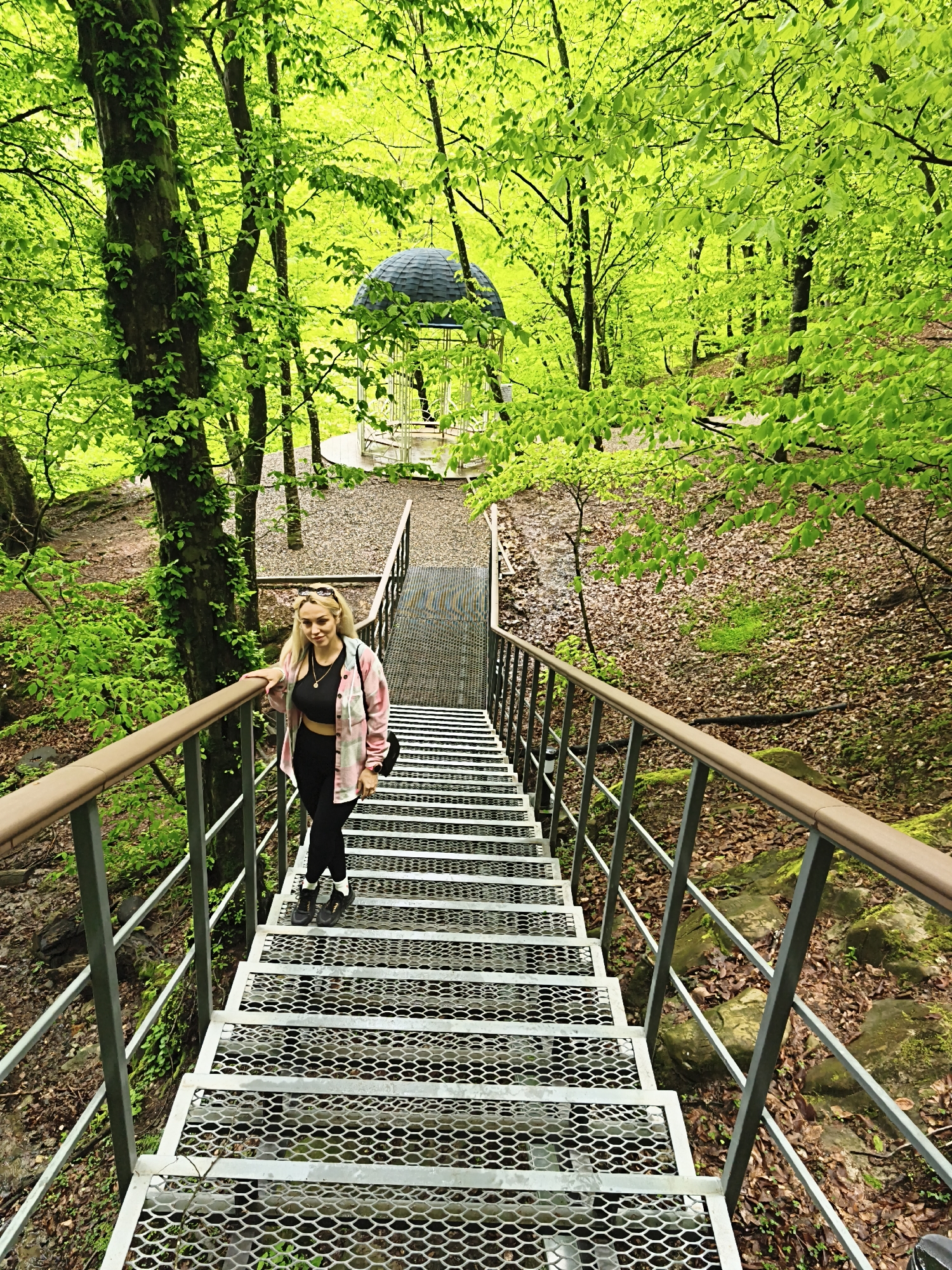 A medium shot shows a woman standing on a metal grate staircase in a forest. The woman has long blonde hair and wears a black crop top, black leggings, a pink and white plaid shirt, and black sneakers. Sunglasses are perched on her head. She holds the handrail with her right hand and faces the camera.
The staircase ascends towards a white gazebo with a dark roof in the background. The surrounding environment is a lush forest with vibrant green trees and foliage. The ground on either side of the staircase is covered in leaves and vegetation. The location is in Goryachiy Klyuch, Russia.