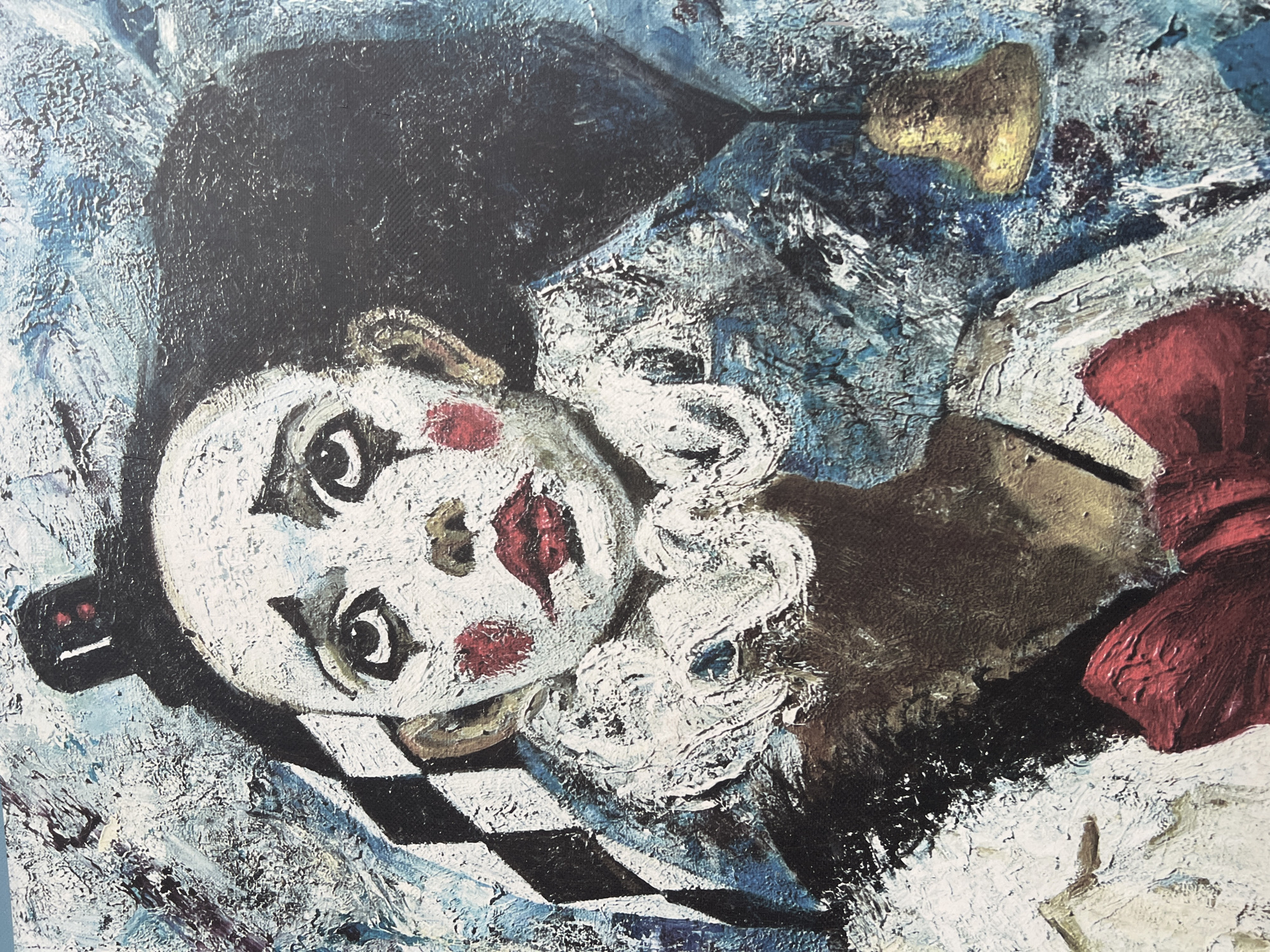 An oil painting depicts a clown. The clown has a white face with red circles on the cheeks and red lips. It has a black hat, a white ruff around its neck, and a large red bow tie. The clown is lying on a checkered surface. The background is a mix of blue and white paint strokes. There is a gold object near the top right. There is no evidence connecting this image to Yerevan, Armenia.