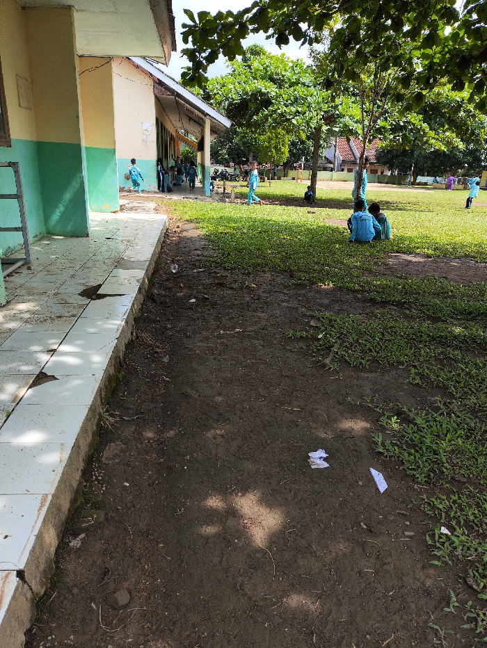 This image captures an outdoor scene at what appears to be a school in Selagai Lingga, Indonesia, on a bright, sunny day. The foreground shows a tiled walkway adjacent to a single-story building with cream-colored walls and a green base. Beyond the walkway is a dirt path, followed by a grassy field dotted with trees that cast shadows.
Several children, primarily boys, are visible, all wearing light blue and white uniforms, typical of elementary students in Indonesia. On the left side, near the building, a few children are walking or standing. In the mid-ground of the grassy field, a group of three boys is seated, possibly resting or chatting. Other children are scattered across the field, some walking, others further in the background.
The activity appears to be a school break or an outdoor lesson, with children leisurely moving or resting. The bright sunlight indicates it is daytime, likely mid-morning or early afternoon, with clear weather. The presence of large, leafy trees suggests a tropical climate. Small pieces of white paper or debris are visible on the dirt path in the lower right foreground. No discernible text is visible in the image.