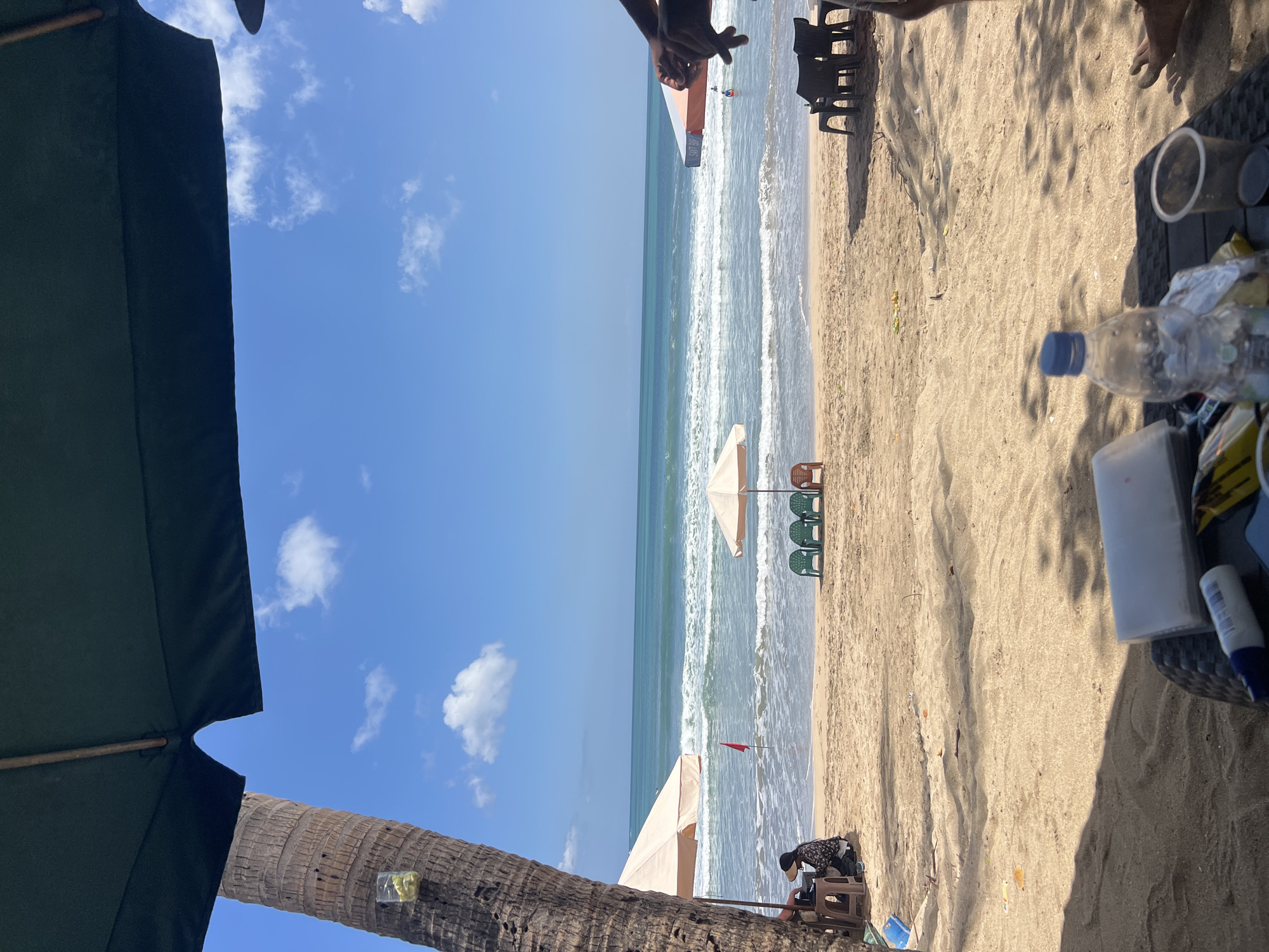 In Kuta, Indonesia, a tranquil beach scene unfolds under a bright blue sky dotted with white clouds. The foreground is dominated by the sandy beach and a table with a plastic water bottle and a cup. A large palm tree trunk is visible on the left, casting a shadow across the sand. In the midground, the ocean stretches to the horizon, with gentle waves lapping at the shore. Beach umbrellas and chairs are scattered on the sand, suggesting a relaxed atmosphere. A few people are visible, one sitting under an umbrella near the palm tree and another person in the water. The scene captures the essence of a typical day at a beach resort.