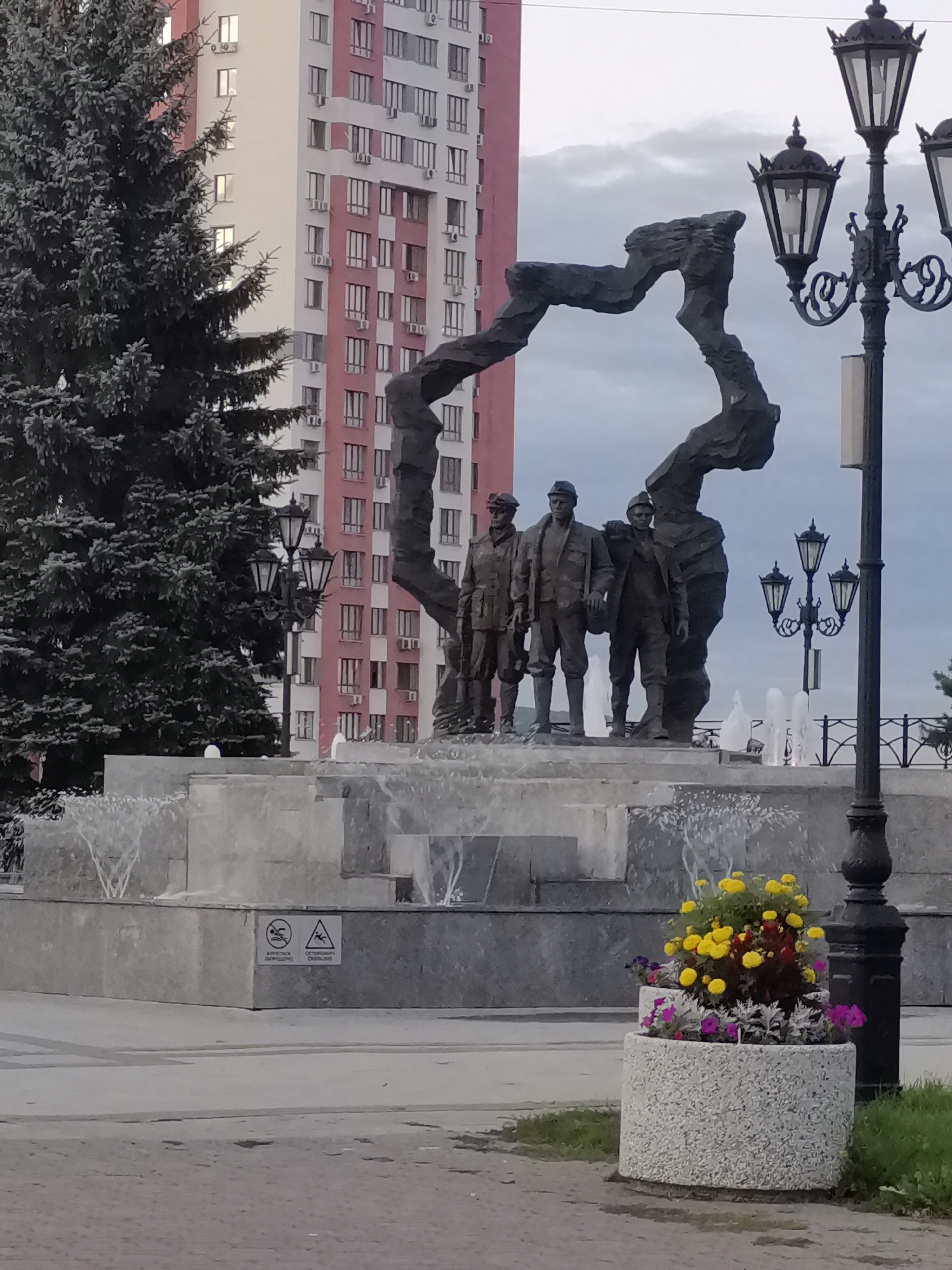 The image depicts a monument in the city of Kemerovo, Russia, situated in front of a tall, modern building with a pink and white facade. The monument features a cluster of three bronze statues of men in historical attire, standing before a large, abstract bronze sculpture with an organic, winding form. A fountain with multiple jets of water is visible behind the statues and sculpture. To the right of the monument, a black, ornate lamppost with three lamps stands prominently, and a concrete planter filled with colorful flowers, including yellow, red, and purple blooms, is positioned in the foreground. The sky is overcast. The scene appears to be an outdoor public space.