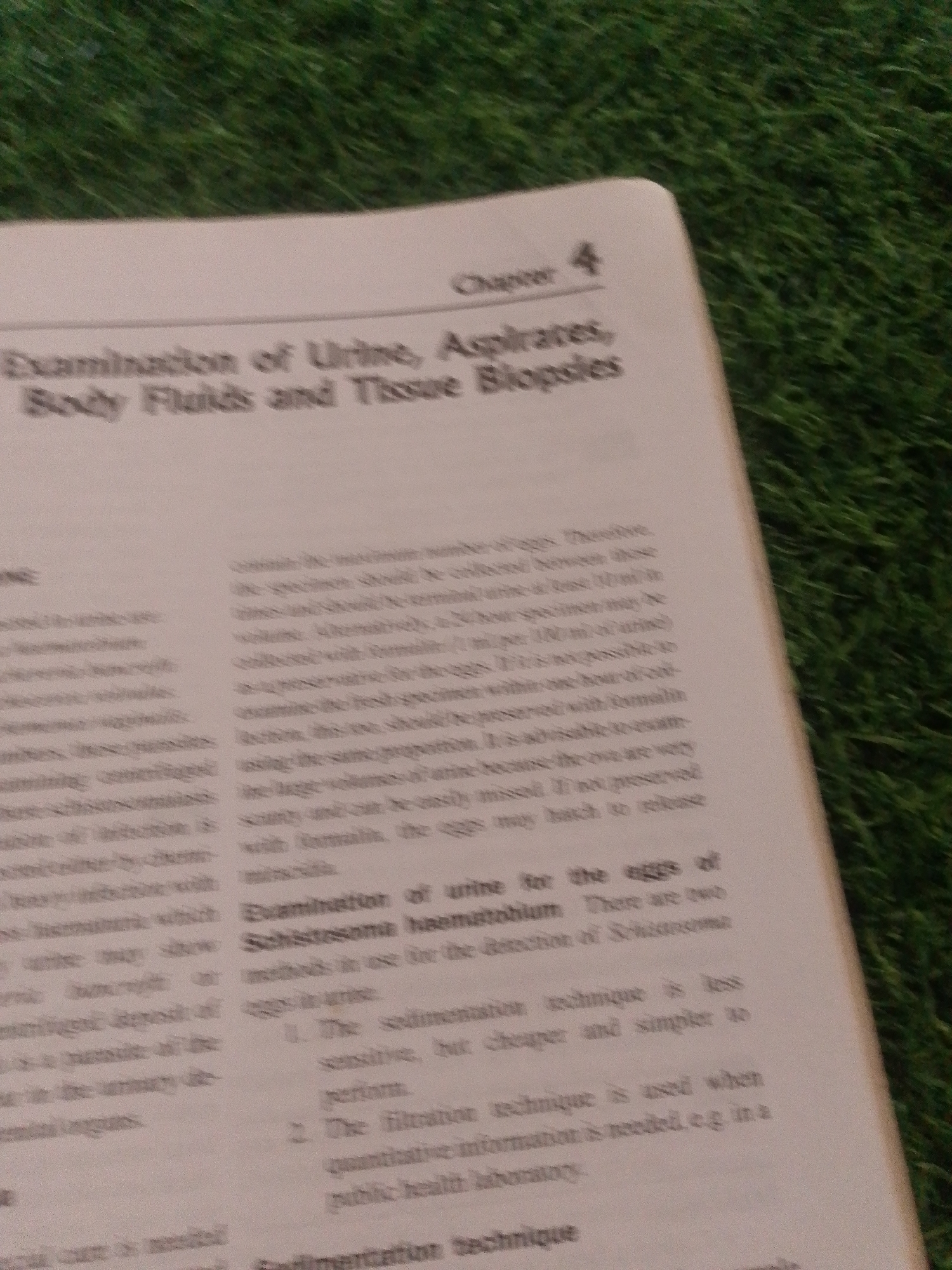 The image displays a close-up, slightly angled shot of an open book resting on a surface that resembles artificial grass. The book's pages are white with printed black text. The left page contains columns of text, some of which are partially visible and blurred. The right page has a prominent title at the top: "Chapter 4". Below this, the title of the section is "Examination of Urine, Aspirates, Body Fluids and Tissue Biopsies". Further down on the right page, there is a subheading "Examination of urine for the eggs of Schistosoma haematobium". The text then enumerates two methods for detection. The overall scene is simple, with the focus solely on the book and its content. There are no people or other objects visible. The lighting appears to be neutral, suggesting an indoor setting, though the artificial grass texture hints at a possible outdoor display or unique decorative choice. The time of day and weather are not discernible. The location context of Jalingo, Nigeria, does not provide any visual cues within the image itself.