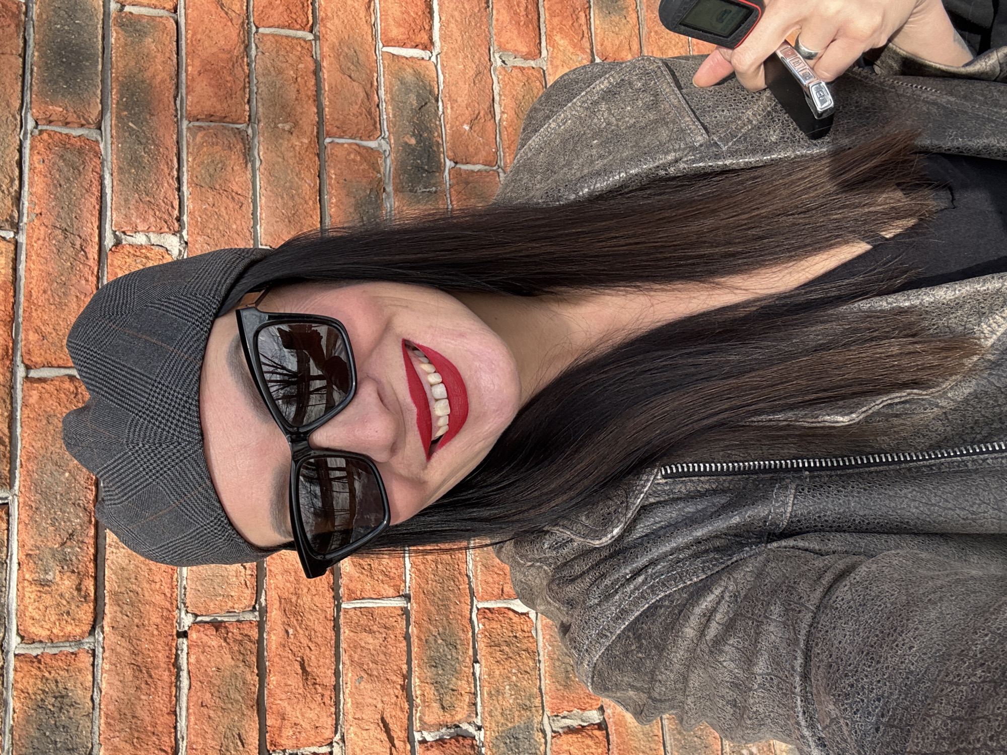A woman is facing the camera wearing a dark gray newsboy cap, black sunglasses, and bright red lipstick. She has long, dark hair. She is wearing a distressed gray leather jacket. In her hand, she holds a car key and remote. The background is a brick wall. The exact location in the city of Chaikovsky, Russia cannot be determined from this image.