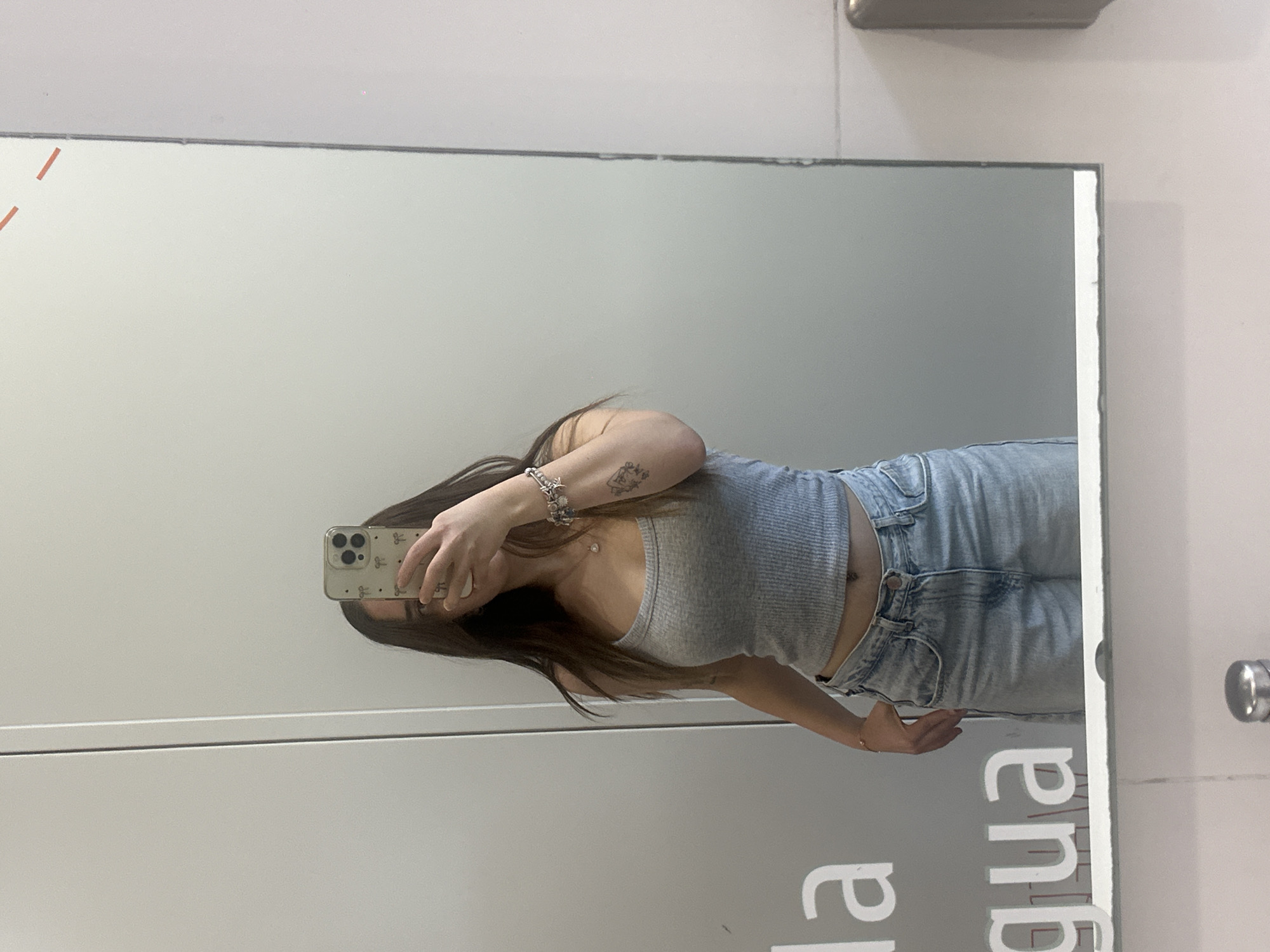 A young woman is captured in a mirror selfie, holding a smartphone that partially obscures her face. She has long, dark hair and is wearing a grey ribbed crop top paired with light blue high-waisted jeans. Accessories include a multi-charm bracelet on her left wrist and a delicate necklace with a small pendant. A tattoo, appearing to be a framed design or symbol, is visible on her upper left arm. Her midriff is exposed, showing a navel piercing.
The selfie is taken in what appears to be an indoor setting, likely a changing room or a room with a large mirror. The walls are plain light grey or white. The mirror itself shows some wear around its edges. In the reflection on the bottom right of the mirror, reversed white text is visible, partially reading "la gua". The woman's smartphone has a light-colored case adorned with a repeating pattern, possibly small bows. The overall impression is casual.