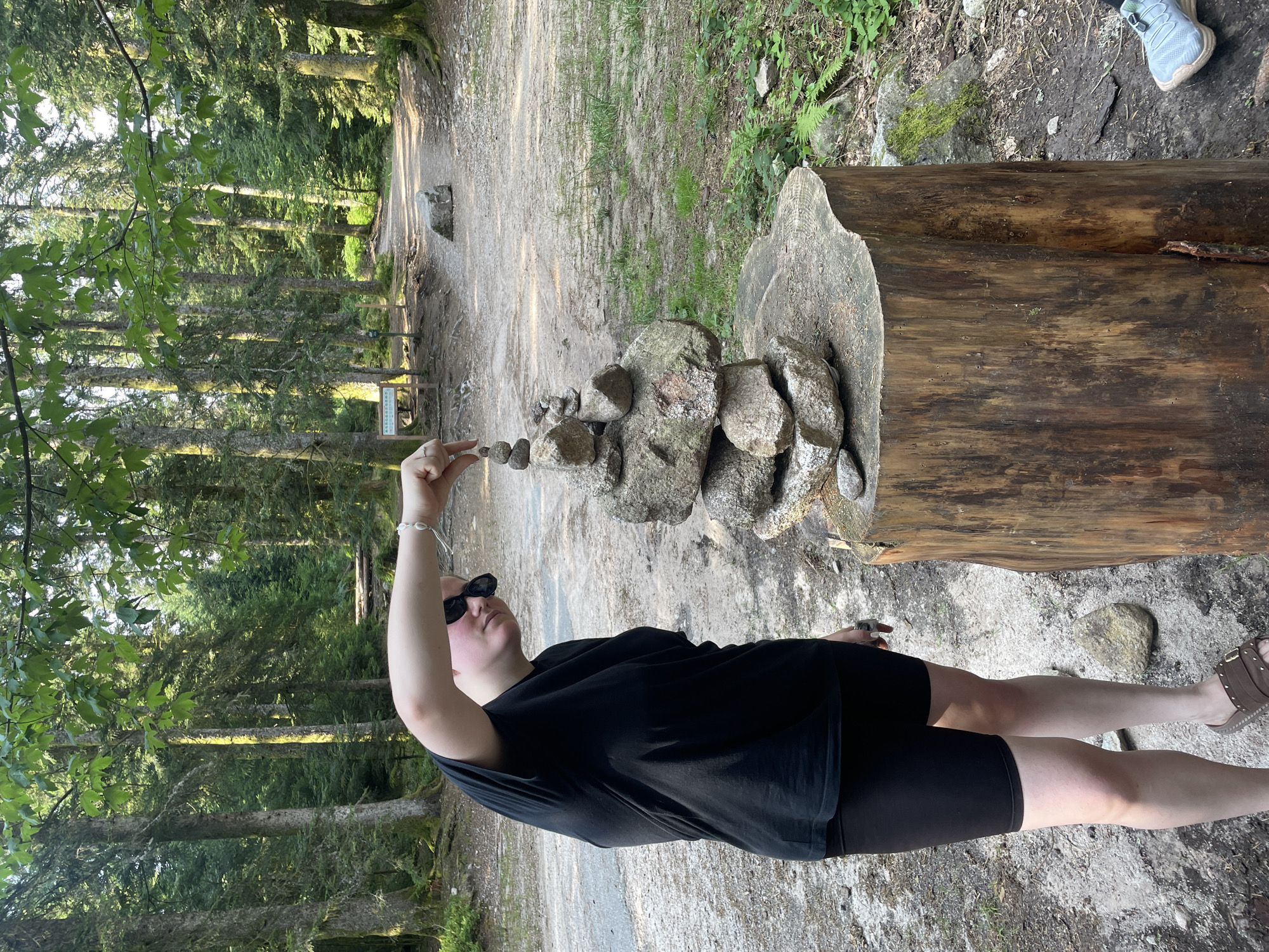 The image shows a person wearing a black t-shirt and black shorts, standing in a forest with a path behind them. The person is balancing a small stack of stones on top of a larger stack of stones, which are placed on a tree stump. The person is wearing sunglasses and has a bracelet on their left wrist. The forest appears to be dense with tall trees. There are other rocks scattered on the ground and a sign in the background. The location is identified as Gérardmer, France.