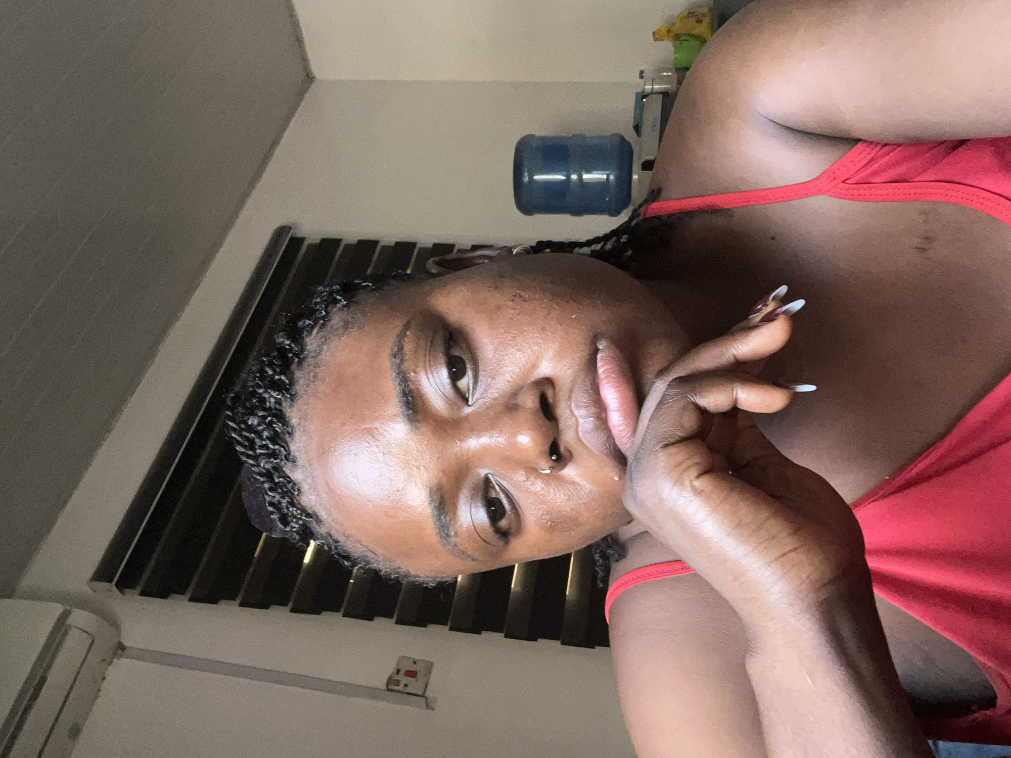 A young Black woman with dark skin and braided hair is seen in a close-up, waist-up shot. She is wearing a red tank top. Her left hand is raised to her chin, with her fingers resting on her lower lip. She has a nose ring and her nails are painted with white tips. Her facial expression appears to be one of contemplation or sadness.
The background is an indoor setting, likely a home or hotel room. To the left, an air conditioning unit is mounted high on the wall. Below it, a light switch with red and white components is visible. Further to the right, a dark-slatted blind covers a window. In the background, a blue water dispenser with a large plastic bottle is situated on a surface, with some other indistinct objects next to it. The walls and ceiling are neutral-colored. The overall lighting suggests it might be daytime, but the room appears to be dimly lit.