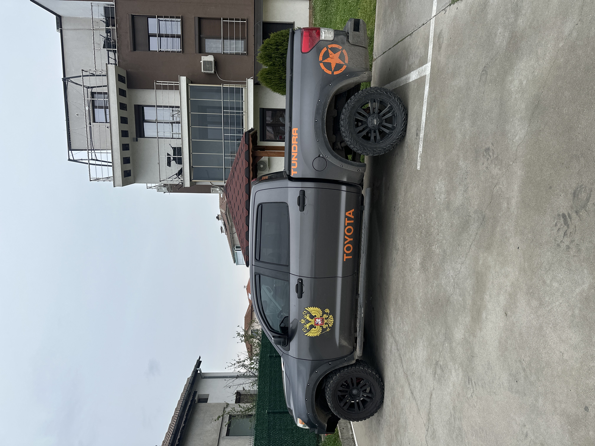 A gray Toyota Tundra pickup truck is parked in a concrete parking space in Svilengrad, Bulgaria. The truck has black aftermarket wheels and is adorned with orange lettering for "TOYOTA" and "TUNDRA." A large orange star is on the rear panel. A gold-colored double-headed eagle decal is on the front door. The parking area is marked with white lines. Buildings are visible in the background. The sky is overcast. The location is likely within a residential or mixed-use area of Svilengrad, Bulgaria.