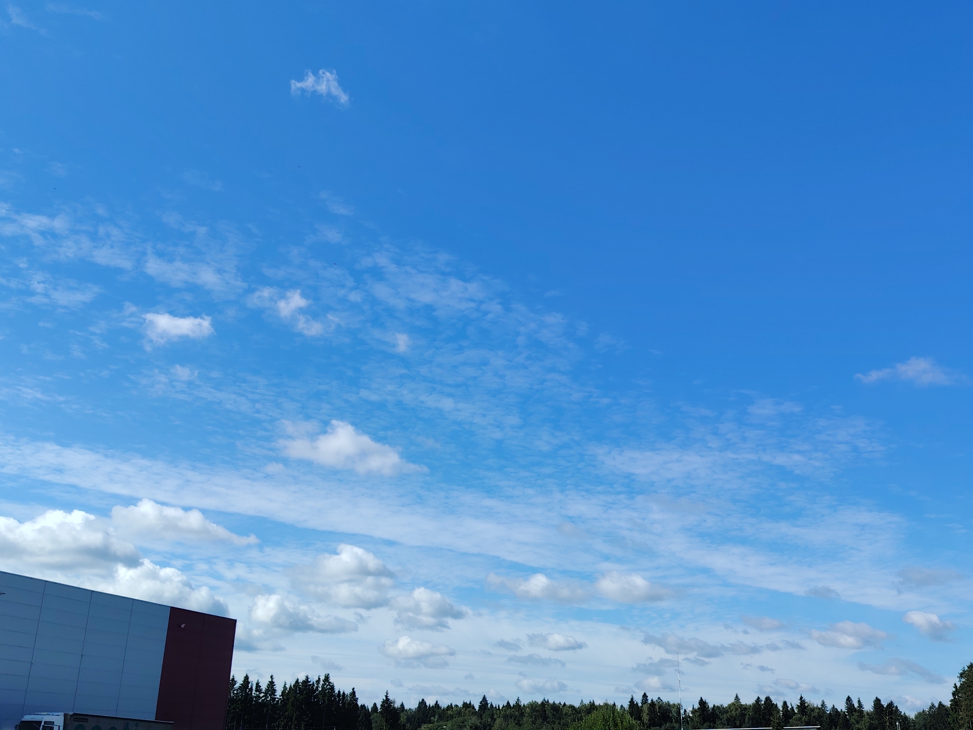 The image depicts a wide shot of a bright blue sky with scattered white clouds. In the lower left corner, a portion of a modern industrial building is visible, featuring a light-colored facade and a dark red vertical section. A white vehicle is parked in front of the building. Below the building and spanning across the bottom of the image is a dense line of dark green coniferous trees. The overall scene suggests an outdoor environment in an industrial or semi-rural area. The location is noted as Khorugvino, Russia.