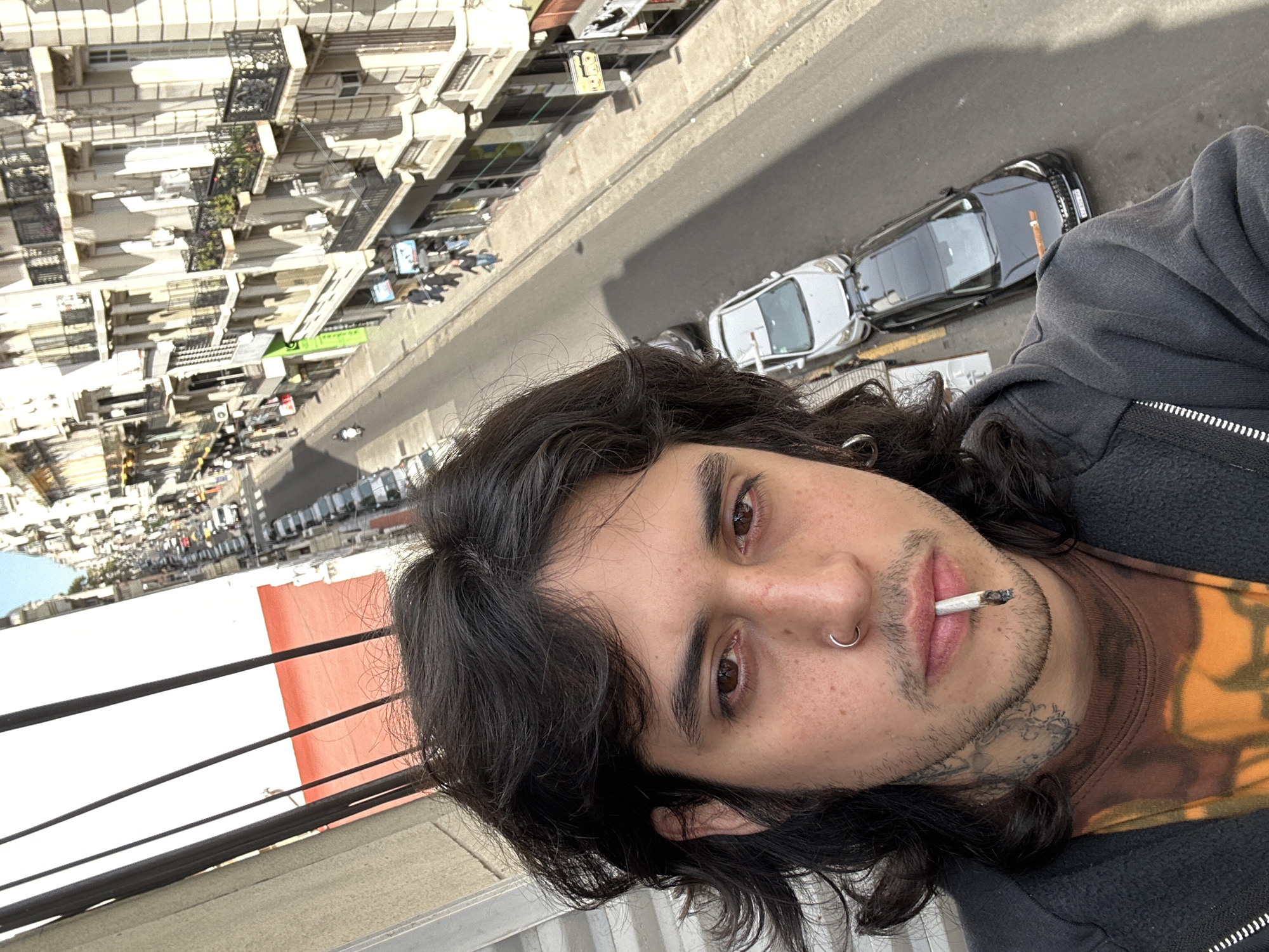 A selfie portrait of a person with long, dark hair, brown eyes, a nose ring, and a cigarette in their mouth. They are wearing a dark-colored hoodie over a patterned t-shirt. The person has visible tattoos on their neck.
The backdrop shows a city street scene with cars parked on the side of the road and tall buildings lining the street. The buildings appear to have balconies and architectural details typical of Buenos Aires. The presence of particular architectural styles like balconies suggests the location is in the city of Buenos Aires, Argentina.