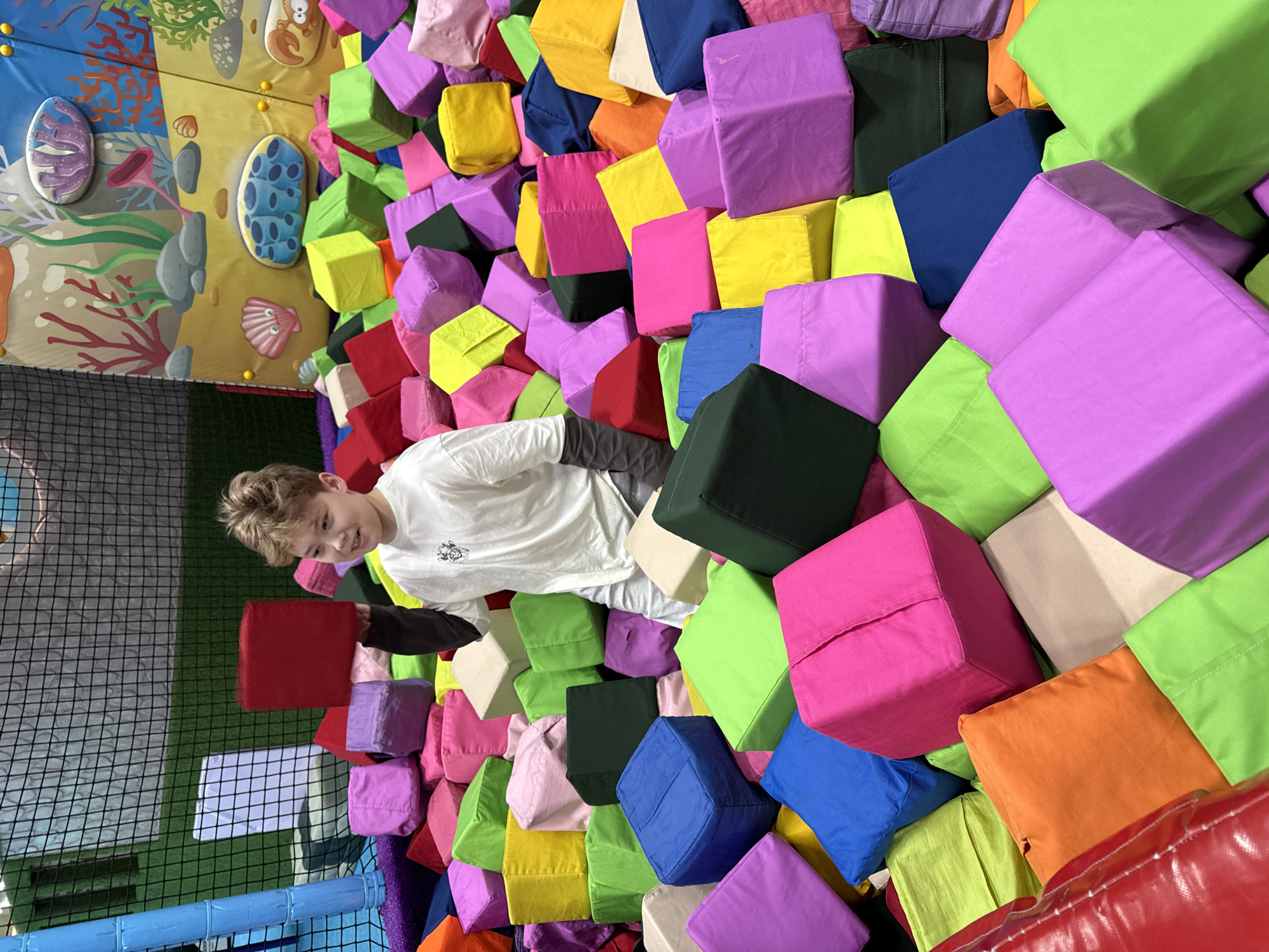 A boy is surrounded by colorful foam blocks in an indoor playground, with a wall mural depicting underwater sea life visible in the background. The playground is likely located in the Перовское сельское поселение, Ukraine.