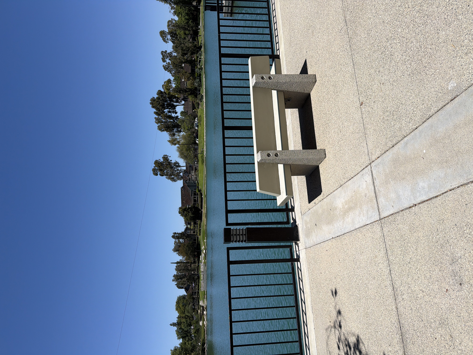 The image shows a tranquil park scene with a clear blue sky and a body of water. In the foreground, a concrete walkway leads to a beige bench situated next to a black metal fence. The fence separates the walkway from the water, which has a vibrant turquoise hue. Across the water, a line of trees and residential buildings can be seen, suggesting a suburban or community setting. The overall impression is one of a peaceful outdoor recreational area. The location is likely a park or public space within the city of Irvine, California, known for its numerous community parks and lakes.