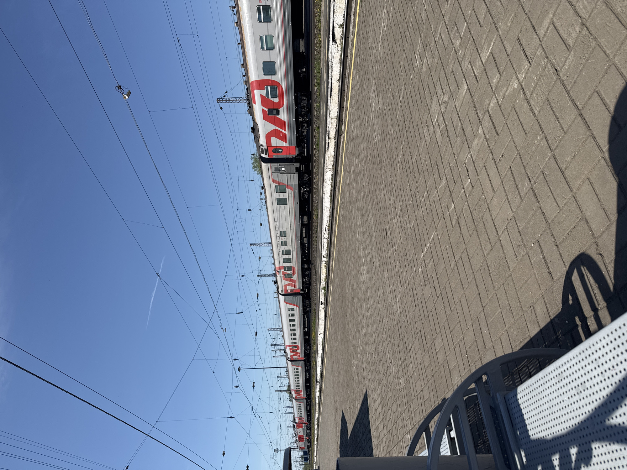 The image depicts a passenger train with "RZD" (Russian Railways) markings at the railway station in Balezino, Russia. The train cars are silver and red. The train is alongside a platform paved with gray brick. Overhead electrical lines run above the train. The sky is clear and blue. Part of a metal bench and its shadow are visible in the foreground on the right side of the image.
