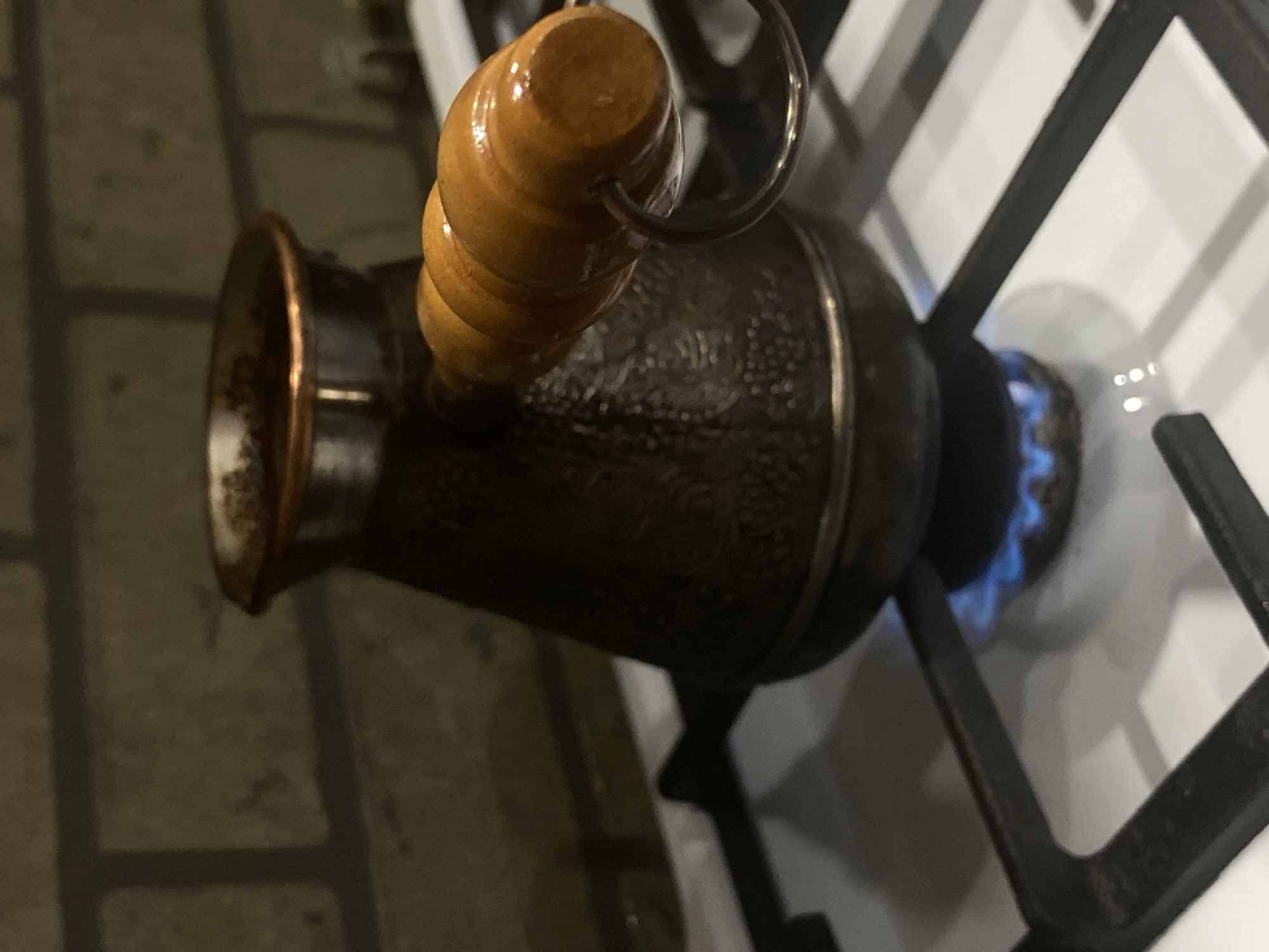 A dark, embossed metallic Cezve, a traditional coffee pot with a light brown wooden handle, is positioned centrally on a white gas stovetop. A blue gas flame emanates from a burner directly beneath the Cezve, indicating active heating. The stovetop features black metal grates. In the background, a dark, textured wall, possibly brickwork, is partially visible under dim lighting. No individuals are present. The specific location within the city of Sochi, Russia, is not ascertainable from this image.