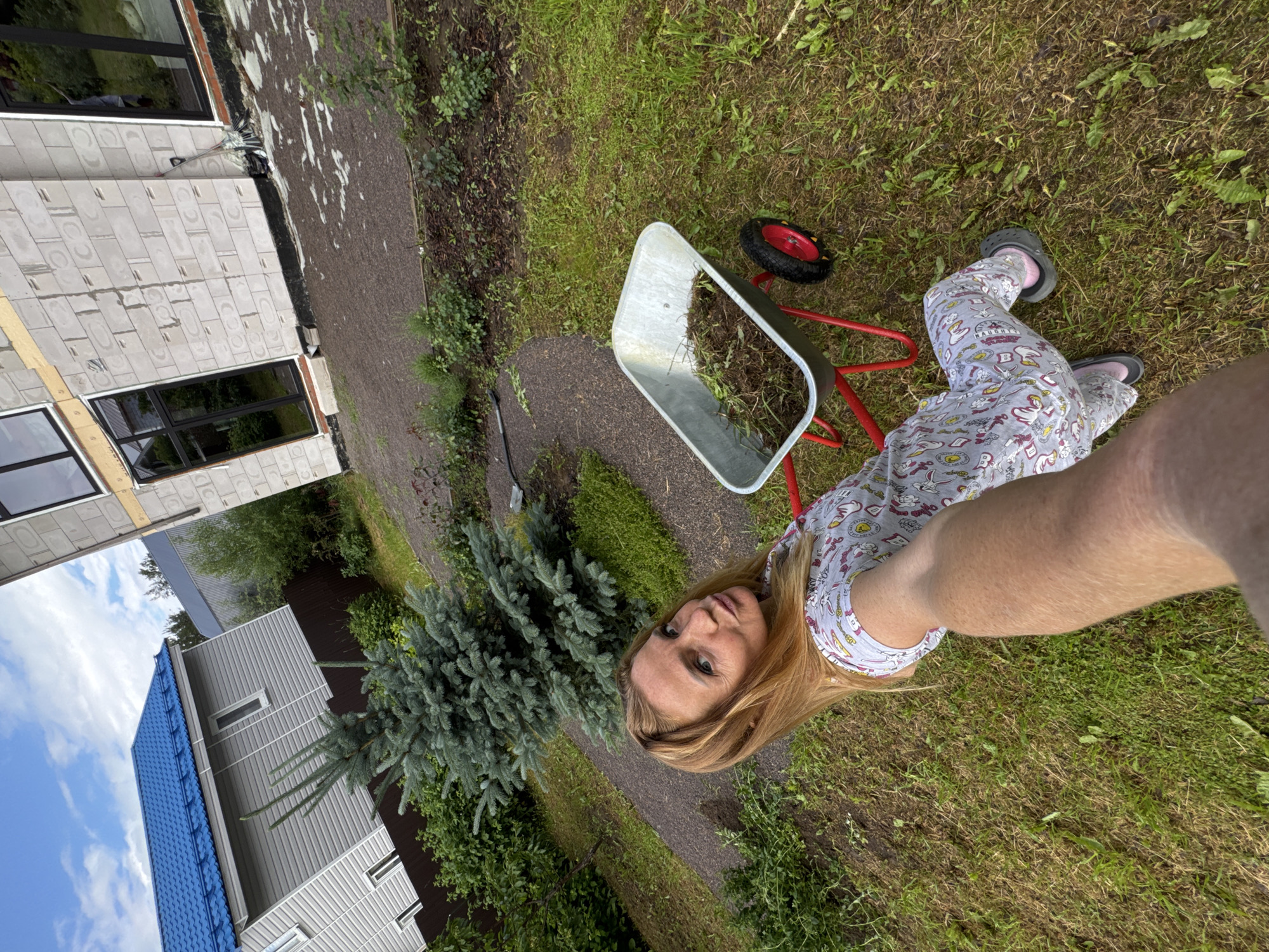 In this full-body, outdoor, slightly downward-angled selfie, a woman with blonde hair and light blue eyes is captured while working in her yard. She is wearing grey pajamas with a cartoon print and dark grey Crocs. The woman is positioned in the foreground, with her face and arm prominently displayed.
Behind her, a wheelbarrow filled with dirt and debris sits on the grass. The wheelbarrow has a red frame and a single red-rimmed wheel. The scene suggests gardening or yard work is in progress.
The background reveals a partially constructed building with white brickwork and dark-framed windows, indicating a residential area under development. Further in the distance, a light grey house with a corrugated metal exterior and a bright blue tiled roof is visible, with lush green trees and bushes surrounding the properties.
The weather appears to be overcast, with the sky showing patches of blue and white clouds. The overall setting is a residential property in Steklyannyi, Russia, with a focus on outdoor activities. The woman's expression is neutral, looking directly at the camera.