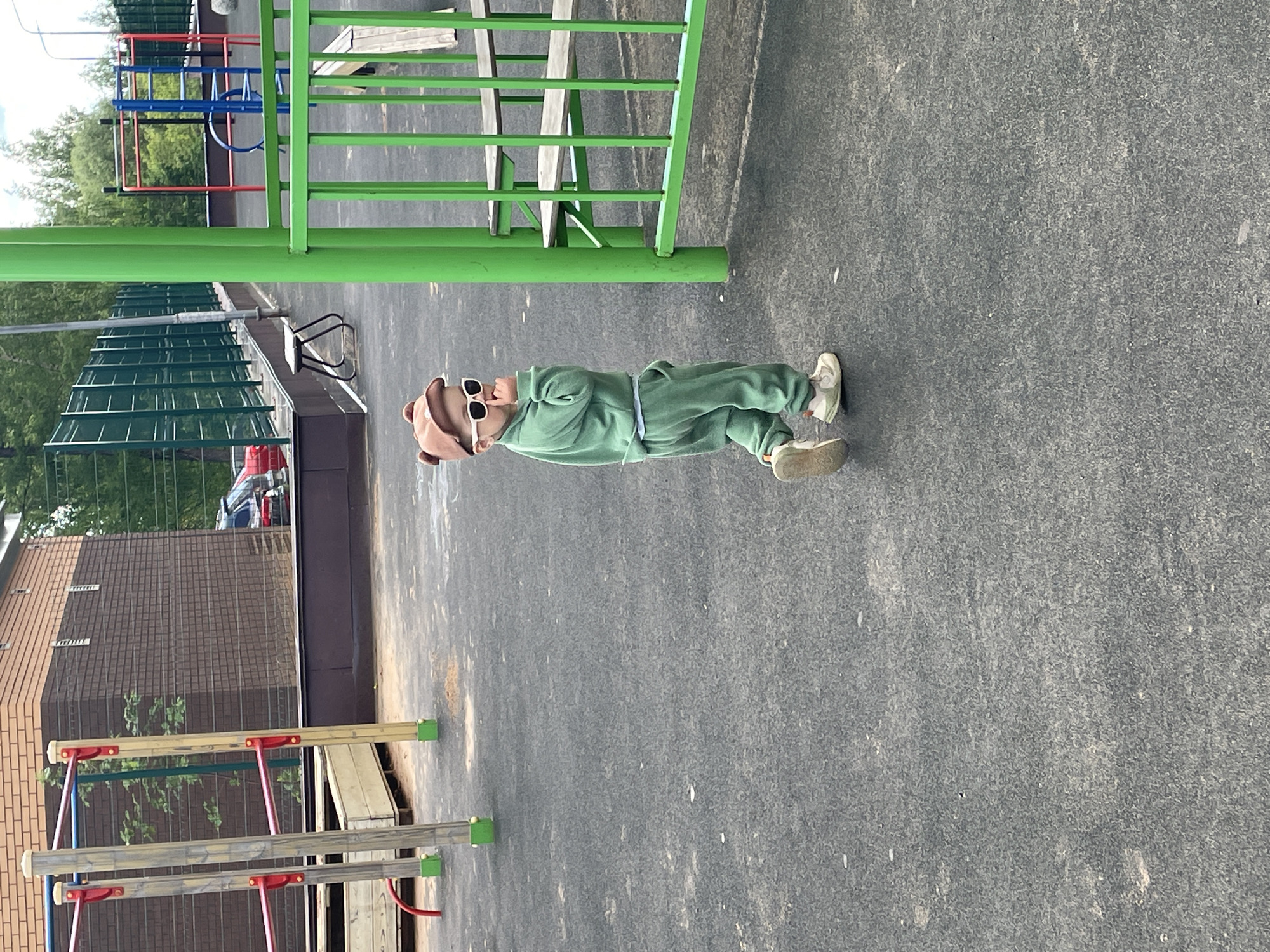 A child in a green sweatsuit, brown cap with ear flaps, and sunglasses is lying on a gray paved surface in what appears to be a playground or recreational area. Playground equipment, including green metal bars and colorful climbing structures, surrounds the child. A building with a brown brick façade and visible drainage pipes is in the background. Based on image metadata, the location is Izhevsk, Russia.