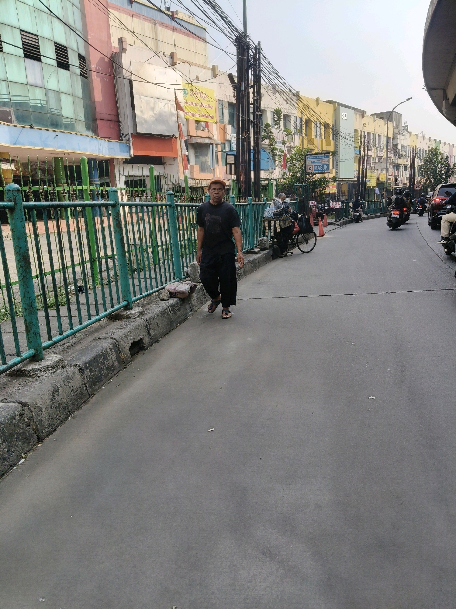 A man wearing a black t-shirt and black pants with a brown cap walks on the right side of a paved street, facing the camera, on a cloudy day in South Tangerang, Indonesia. He is walking along a curb next to a green metal fence. Behind the fence are buildings with colorful facades and signs, including one in yellow that reads "Busana Muslim & Seragam Sekolah" (Muslim Clothing & School Uniforms). Several motorcycles are on the street, some parked and others in motion. A red car is also visible on the right side of the street. The environment appears to be a commercial or mixed-use urban area.