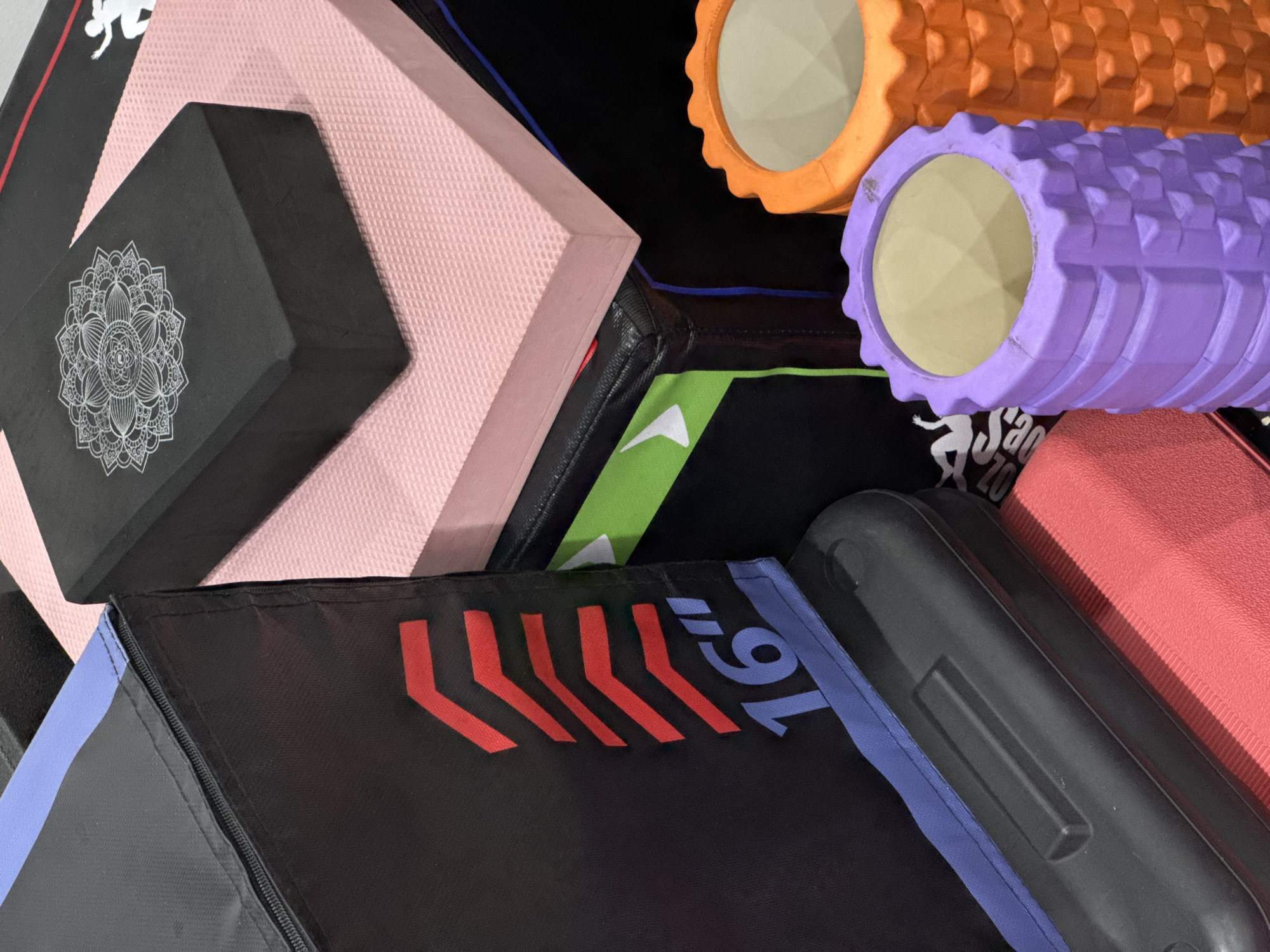 The image shows a collection of fitness equipment, including two foam rollers (one purple, one orange), a pink yoga mat, a black yoga block with a mandala design, and a black fitness box with red chevron markings and the number "16" in blue. A black rectangular object, possibly a mat or another piece of equipment, is partially visible in the foreground, resting on a red mat. The items are arranged in a somewhat cluttered manner, suggesting they are stored or prepared for use. The lighting is moderate, creating shadows that define the shapes of the objects. No people are present in the image. The background is dark and out of focus, providing no contextual information. No specific location within Nam Nha Trang Ward, Vietnam, can be determined from the visual evidence.