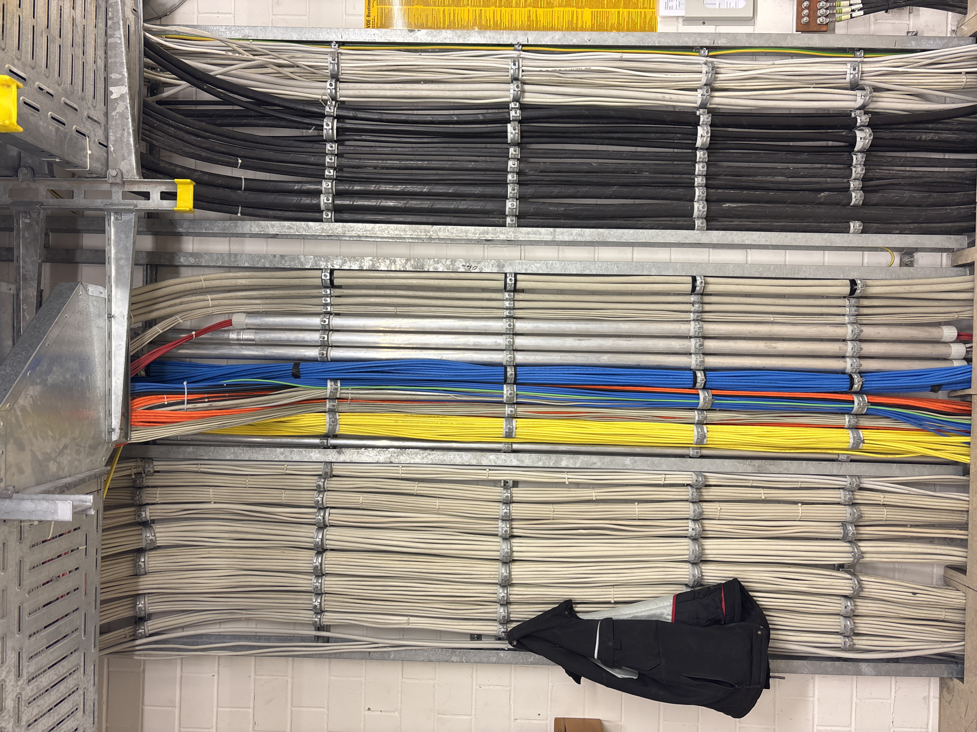 The image shows a section of a utility room with a large number of organized electrical cables. The cables are bundled and secured with metal clamps to a metal frame attached to a brick wall. There are multiple rows of cables, predominantly beige and black, with some colorful cables (yellow, orange, blue, red) visible in the center. A black jacket is hanging on the left side of the cable bundles. Above the cables, metal conduit and cable trays are visible. On the right side of the image, a yellow sign with German text is mounted on the wall, and further down, electrical junction boxes and connectors are present. The overall environment suggests a technical or industrial setting. The location of Mannheim, Germany is not discernible from the image itself.